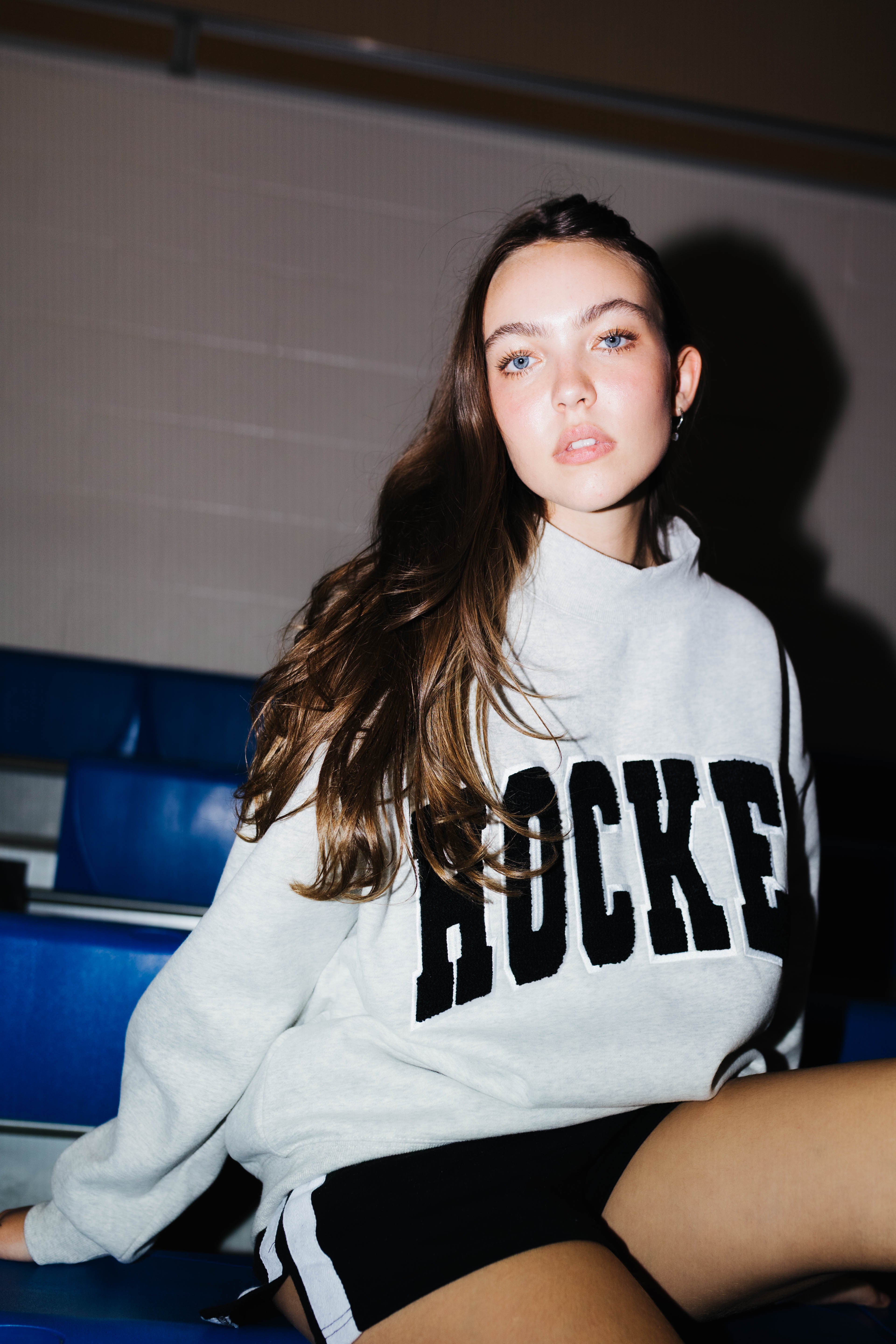 A woman sitting on blue stadium seating wearing a light gray sweatshirt with 'HOCKEY' printed in black across the chest, paired with black shorts.