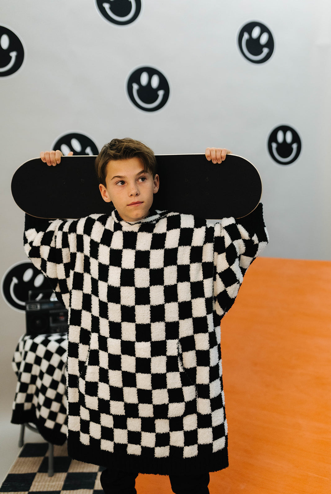 Person wearing a black and white checkered outfit holding a skateboard against a wall with smiley face designs.