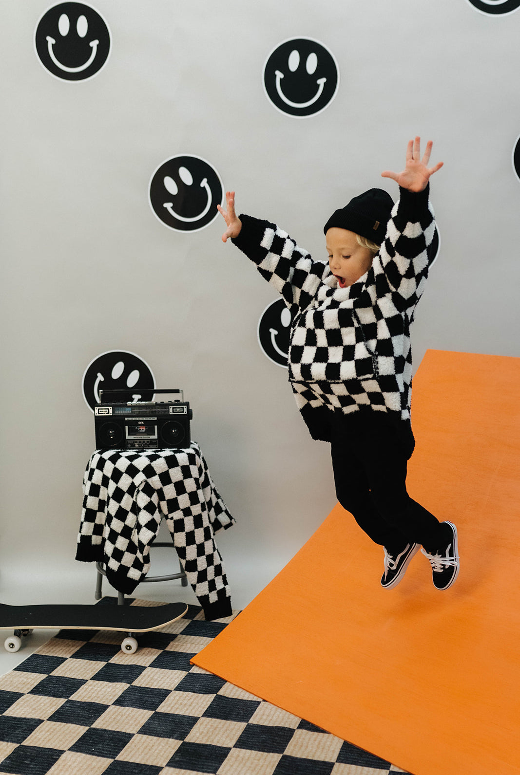Person in a checkered wearable jumping on an orange mat with smiley face decorations on the wall.