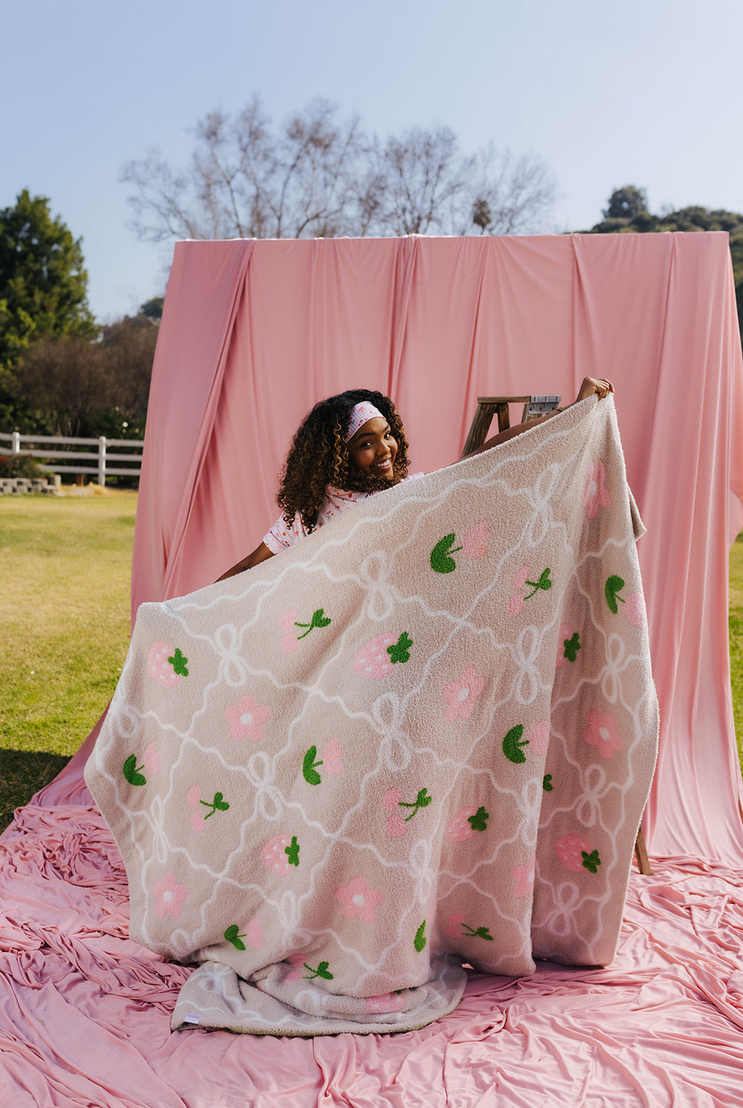 Woman holding a sand blanket with pink and green patterns in front of a pink backdrop outdoors.