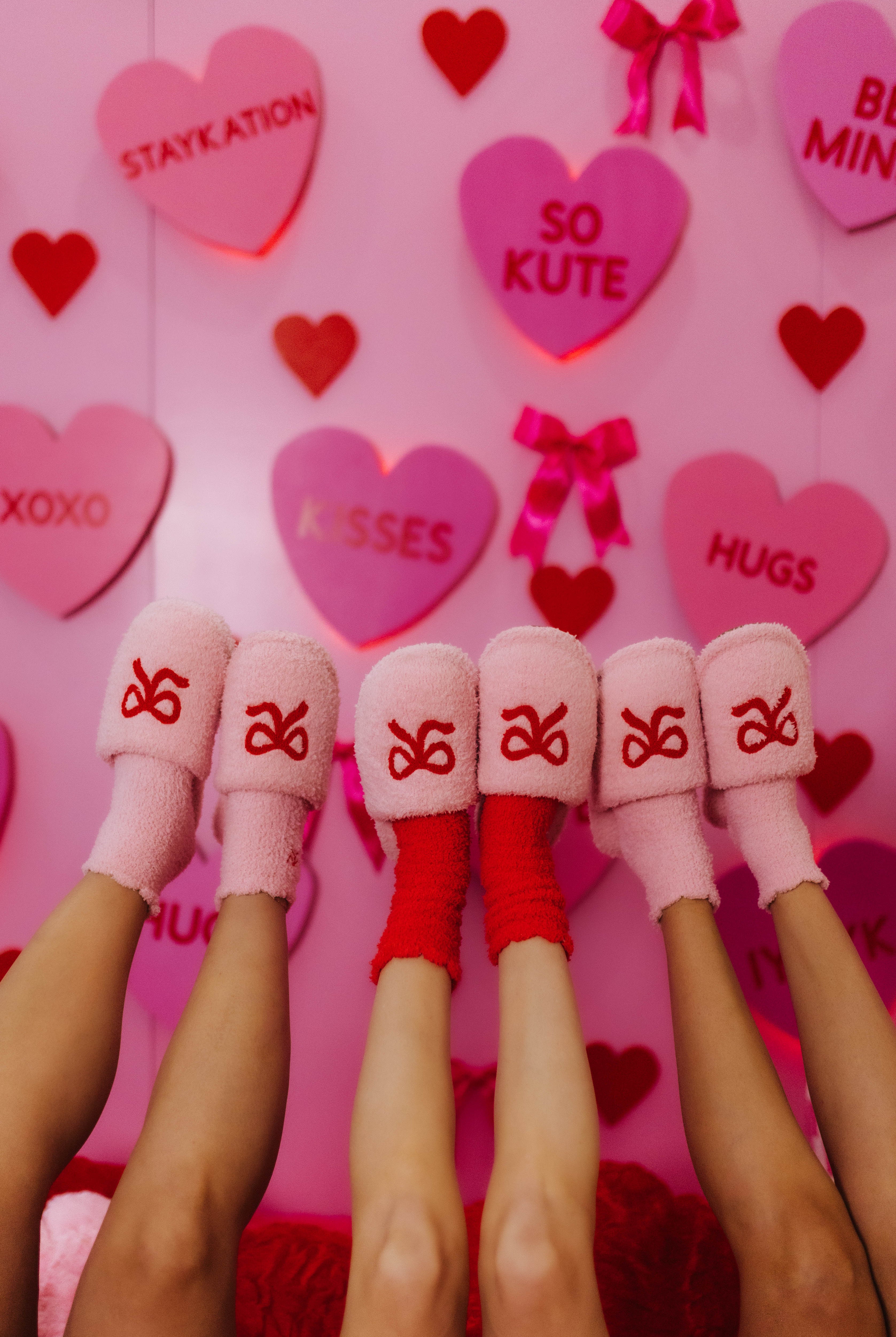 Pink and red socks with slippers in front of a pink background with heart-shaped text and decorations.