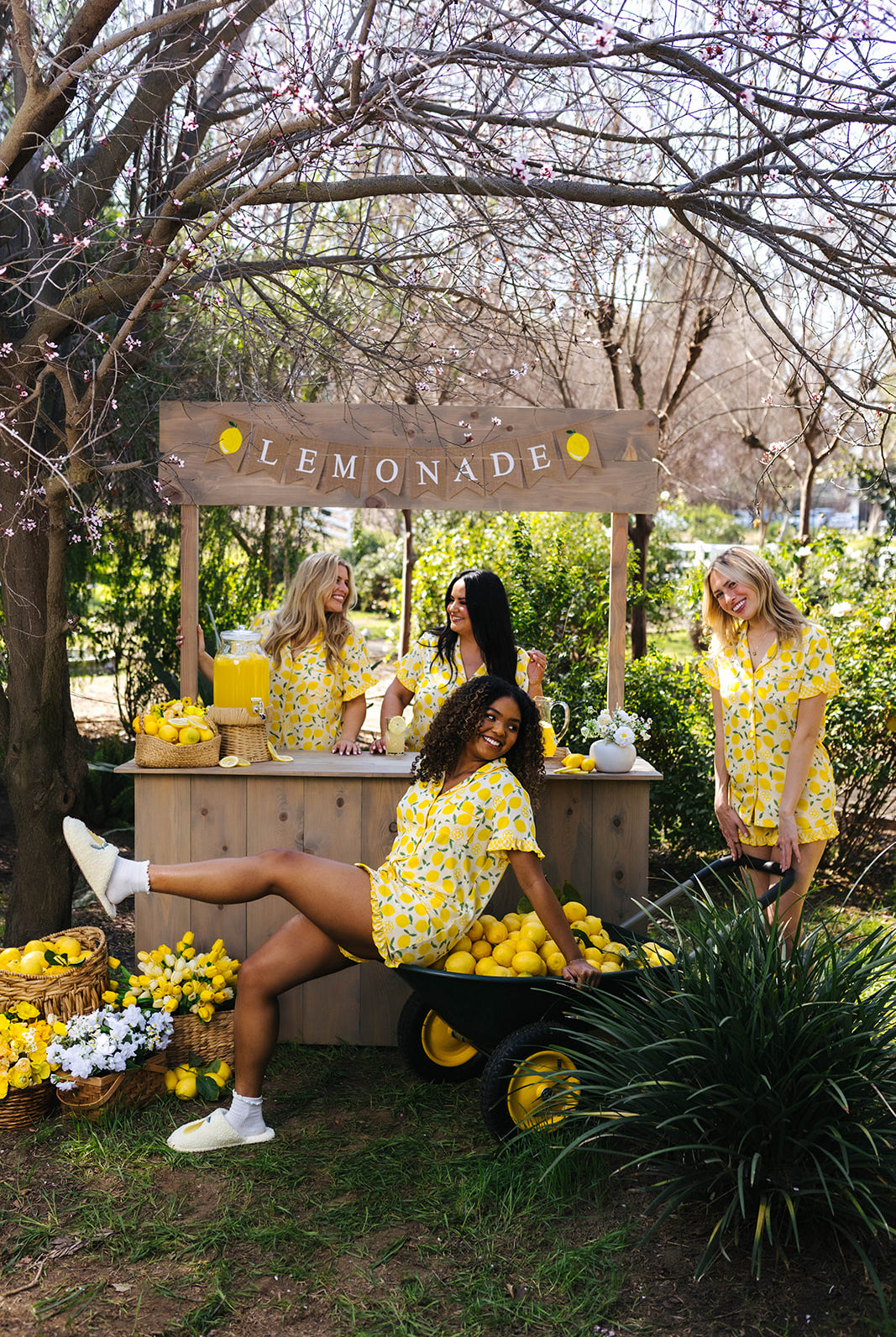 Four women in yellow pjs standing around a lemonade stand with lemonade and lemons in an outdoor setting.