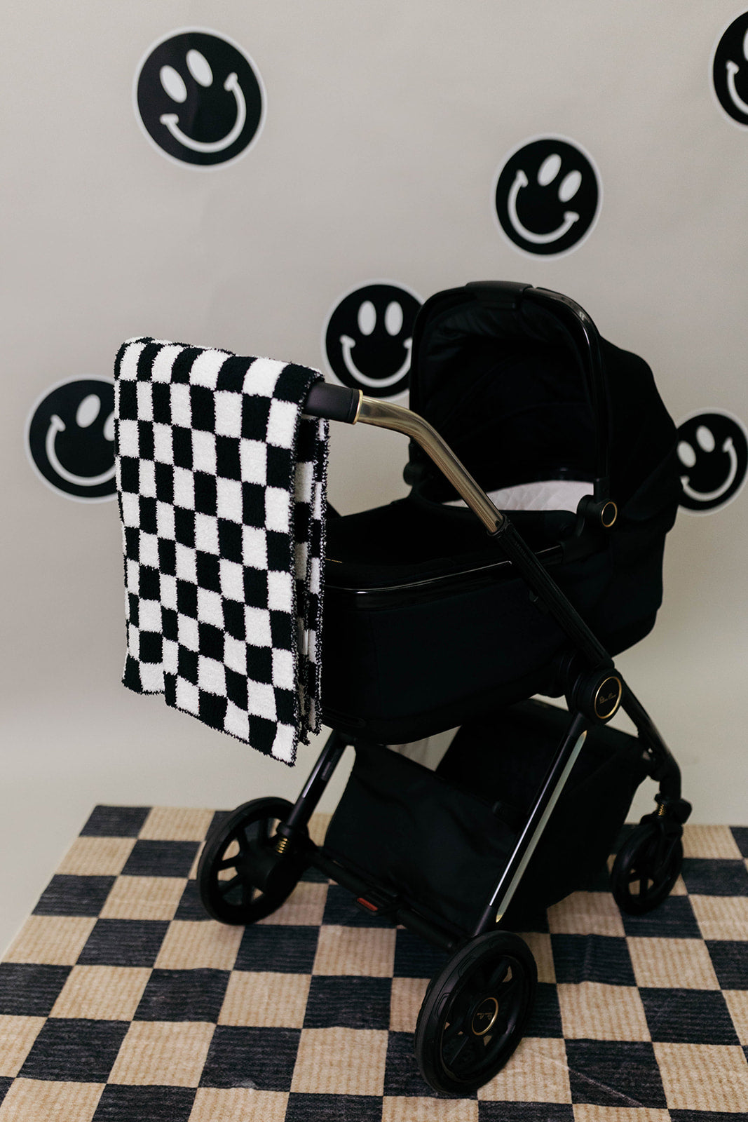 Black stroller with a checkered blanket on a checkered floor against a wall with smiley face designs.