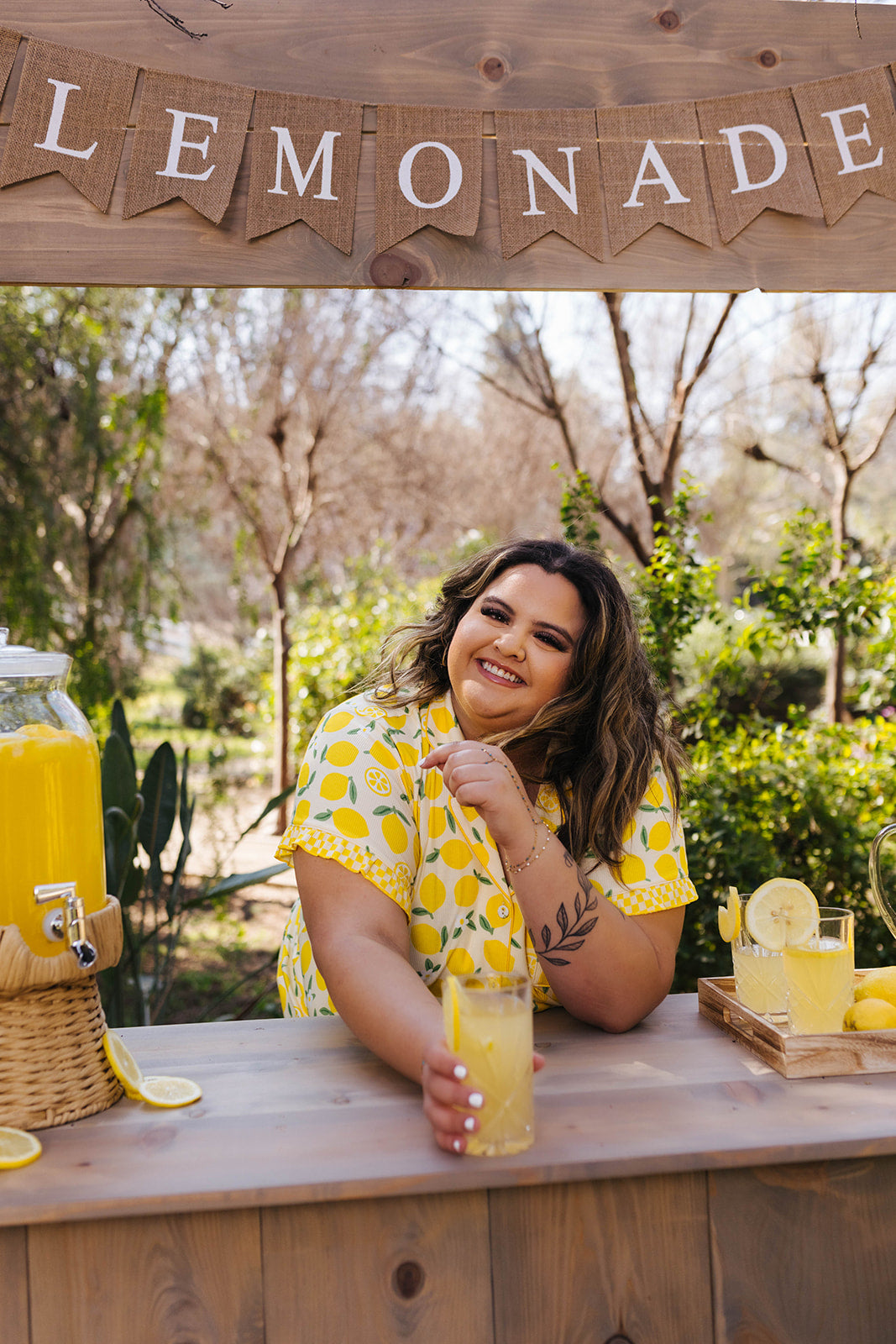 Woman sitting at a lemonade stand with lemonade and lemons, surrounded by trees.