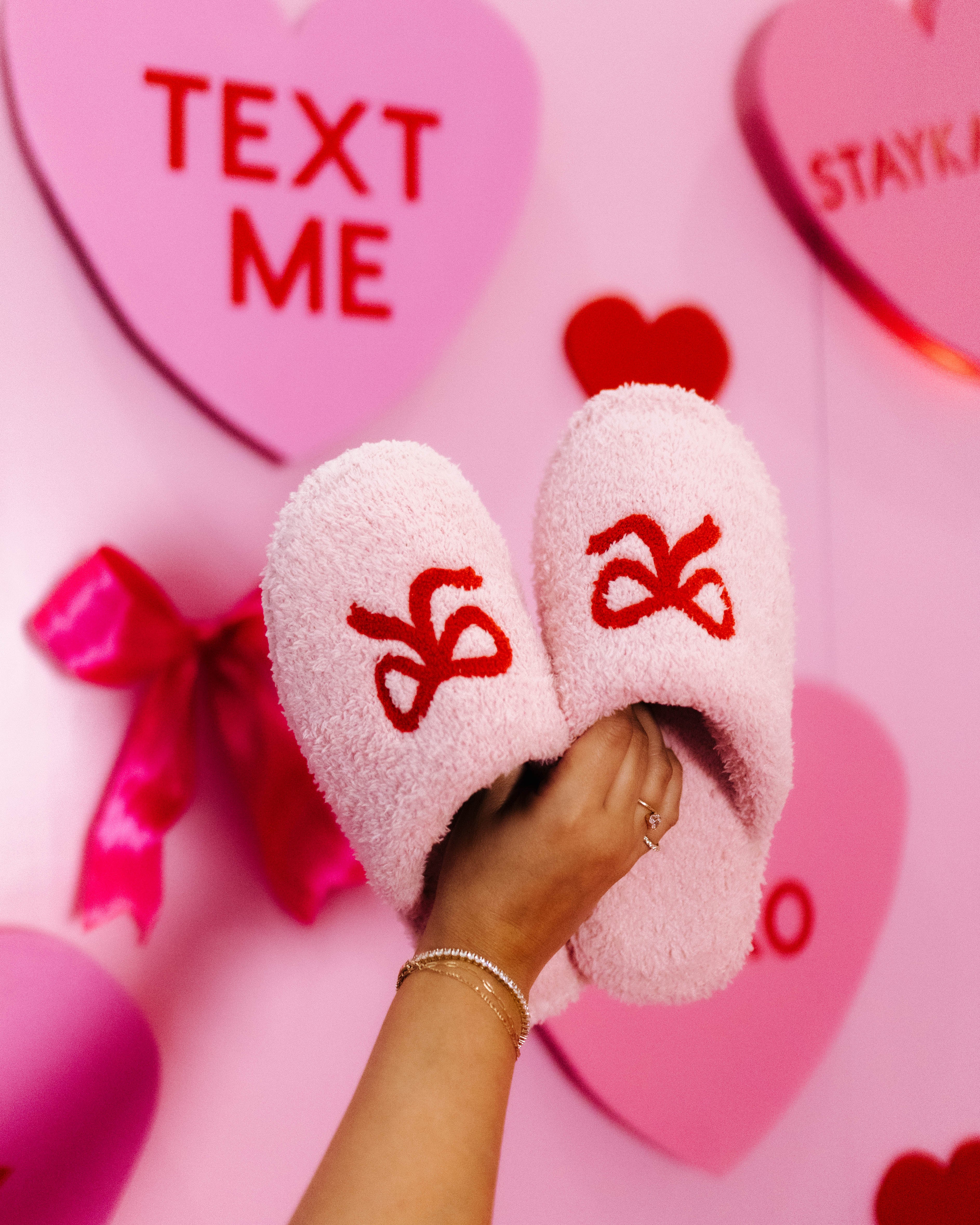 Pink fuzzy slippers with red bow design on a pink background with hearts and text.