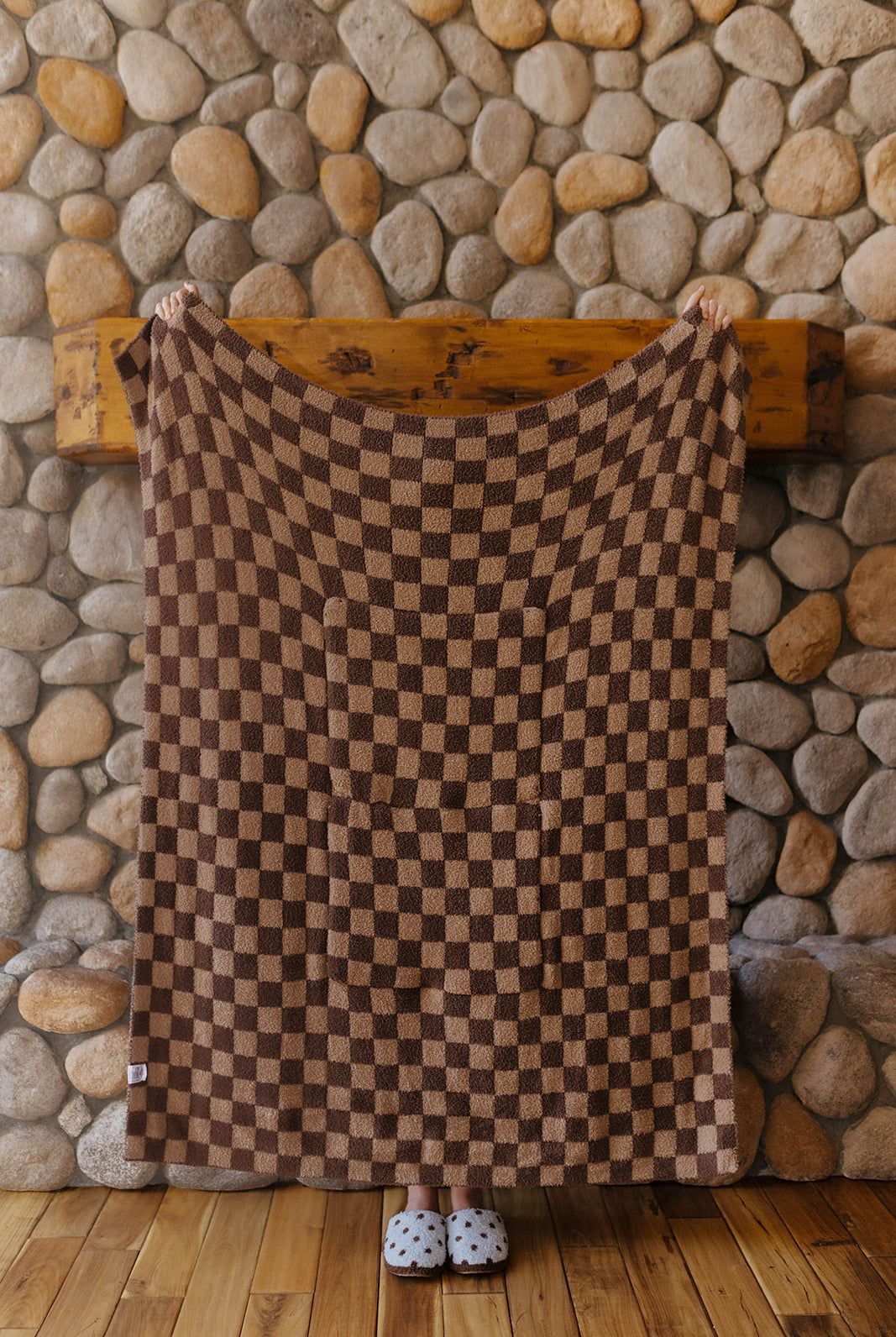 Brown check quillow opened into a blanket in front of a stone fireplace.