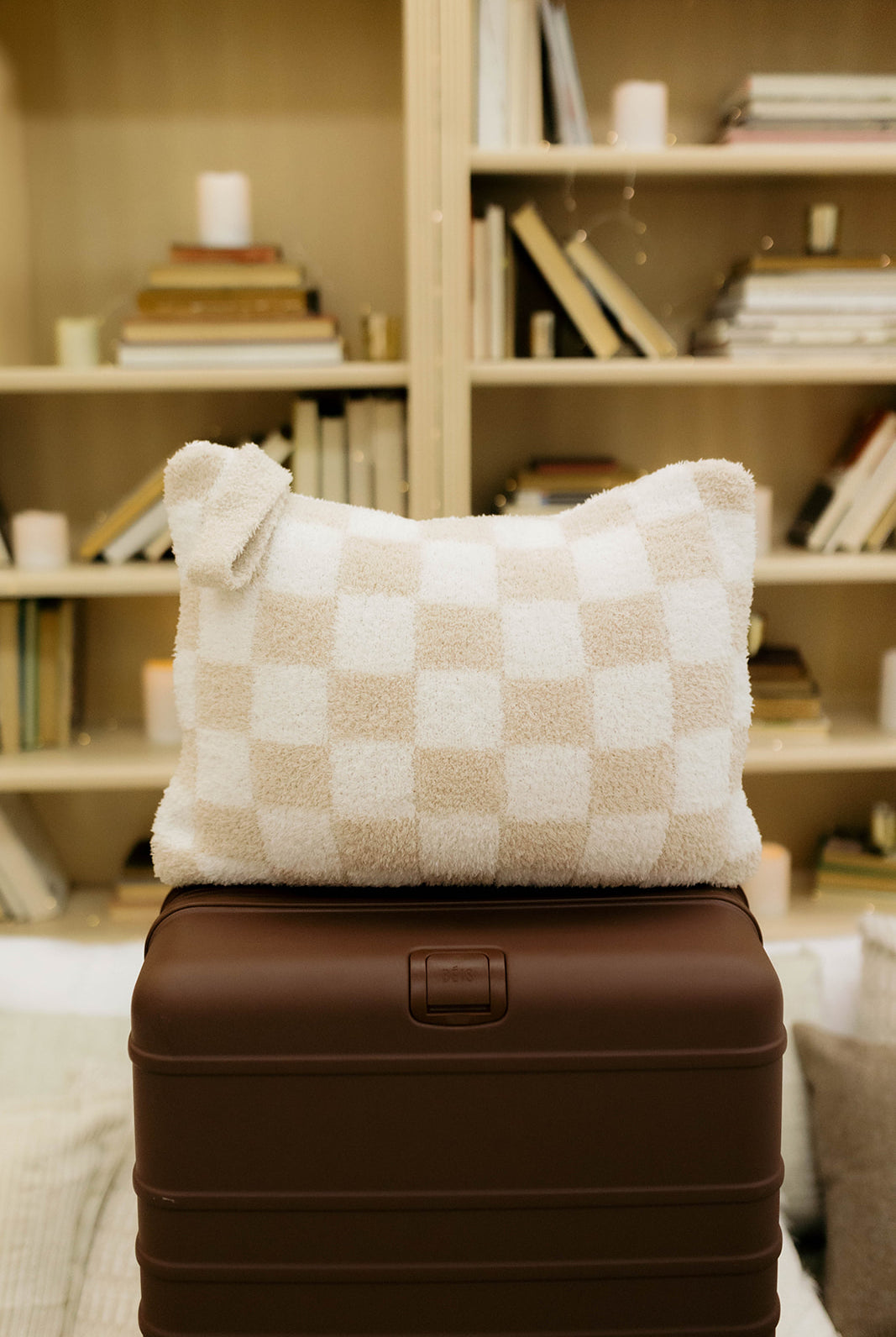 Checkered pillow on a brown suitcase with a bookshelf in the background