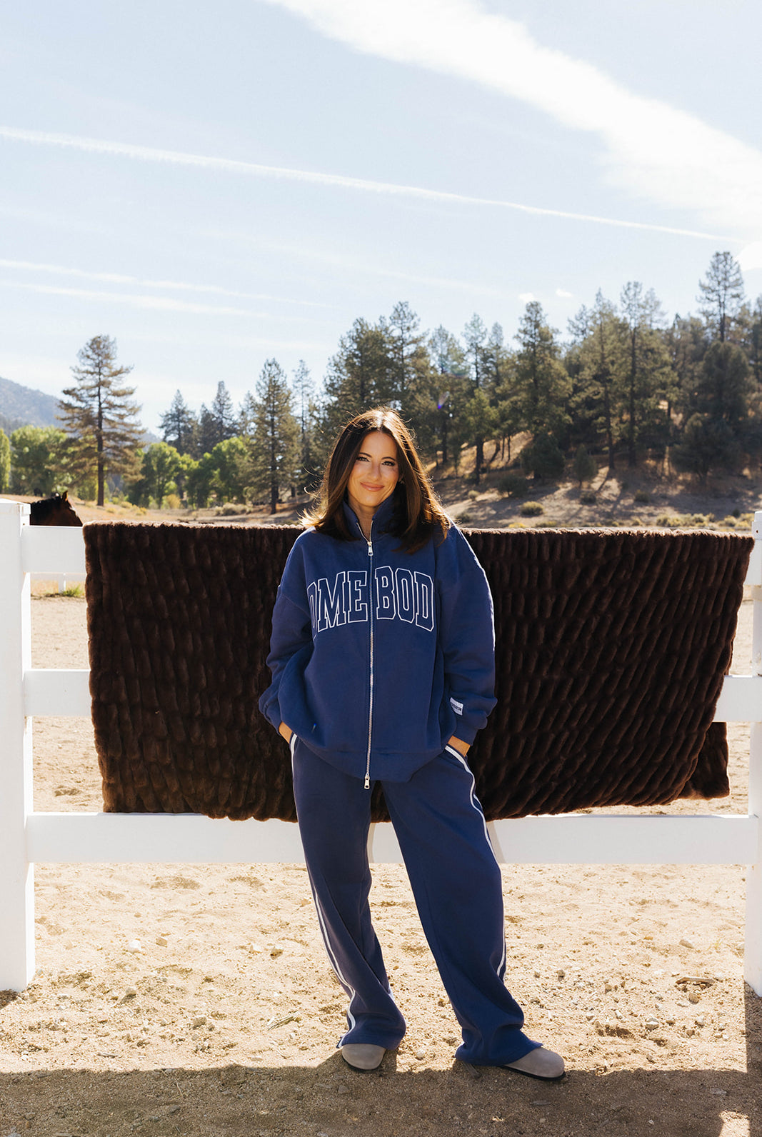 Full length front view of the blue Homebody zip sweatshirt and side stripe sweatpants beside a fence.