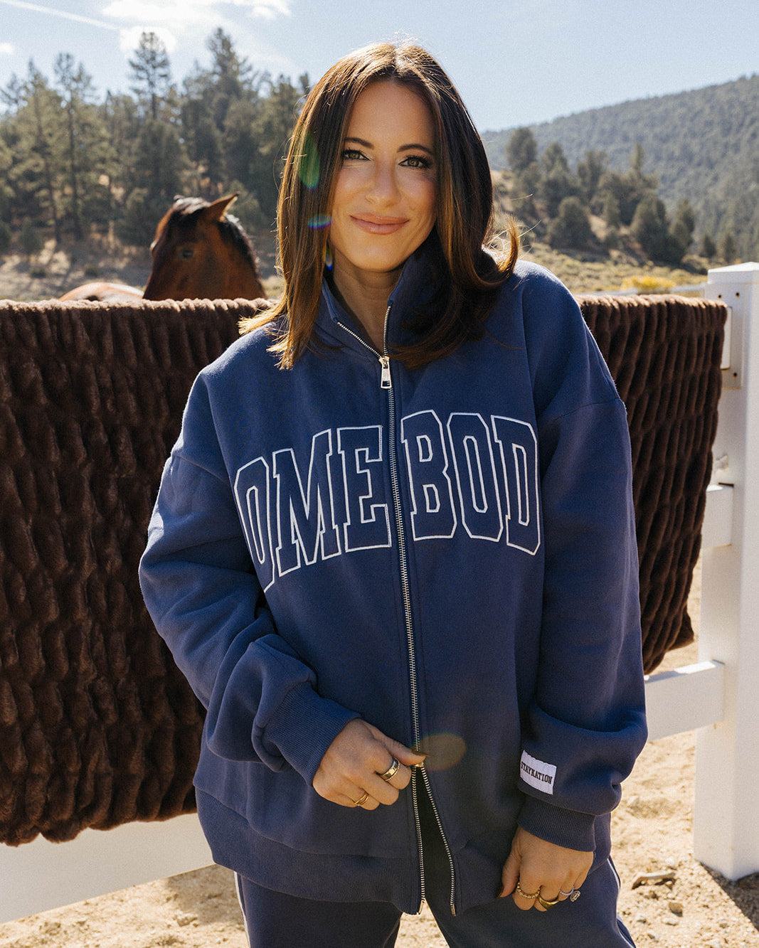 Model wearing the blue Homebody zip sweatshirt by a white fence.