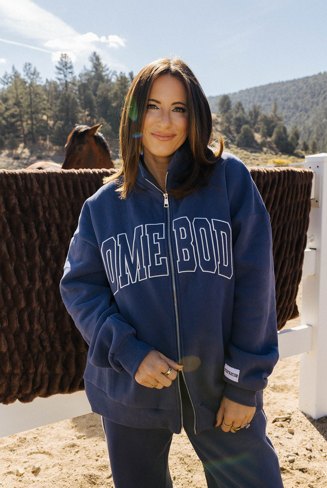 Model wearing the blue Homebody zip sweatshirt by a white fence.