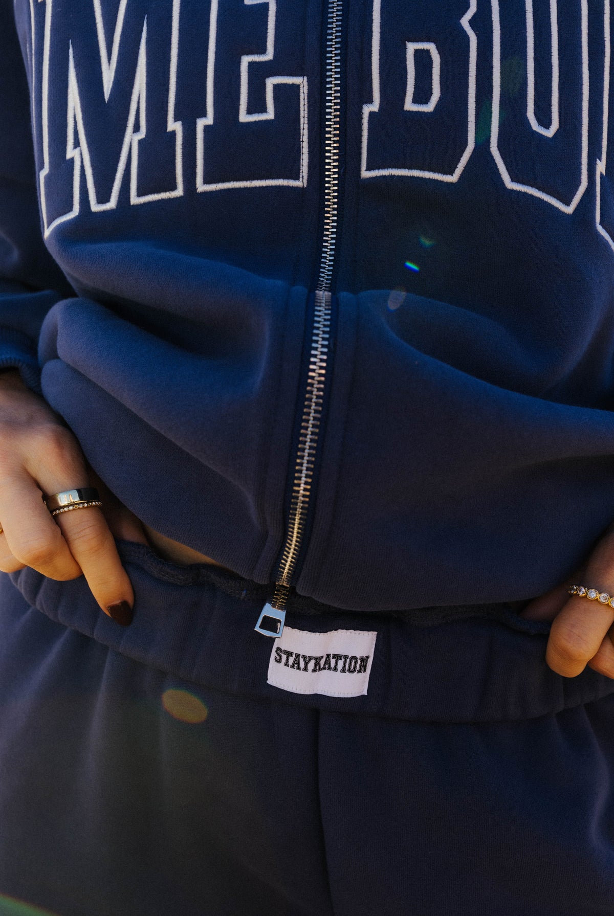Detail of the navy Homebody zip sweatshirt and pants showing the zipper, white piping, and Staykation waistband label.