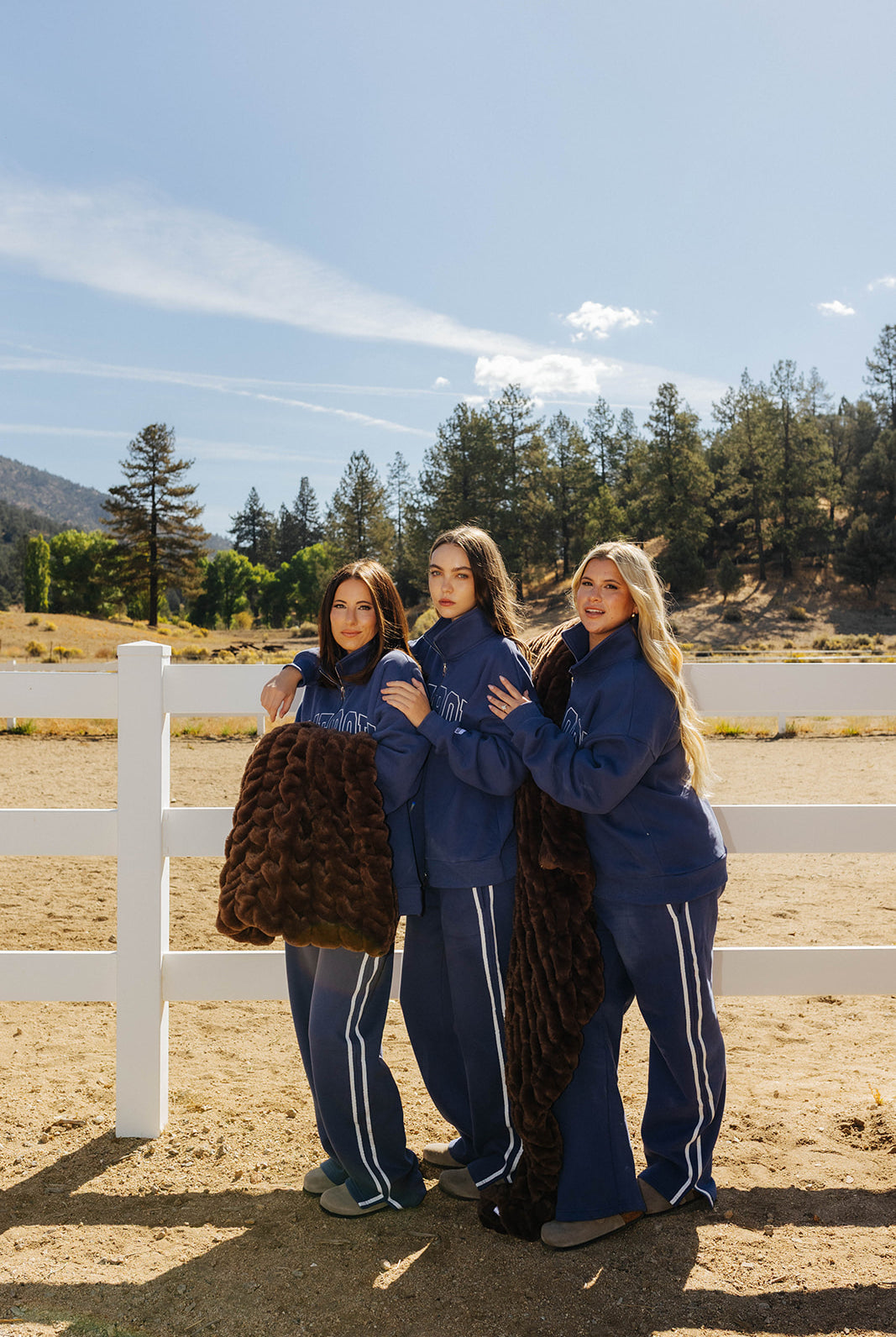 Three models wearing navy Homebody sets and holding a chocolate brown faux fur blanket beside a white fence.