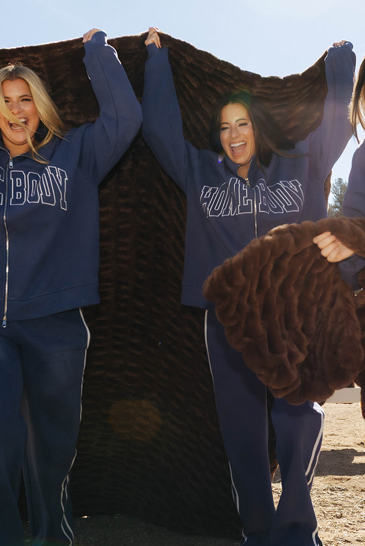 Models holding chocolate brown faux fur blankets open beside a white fence.