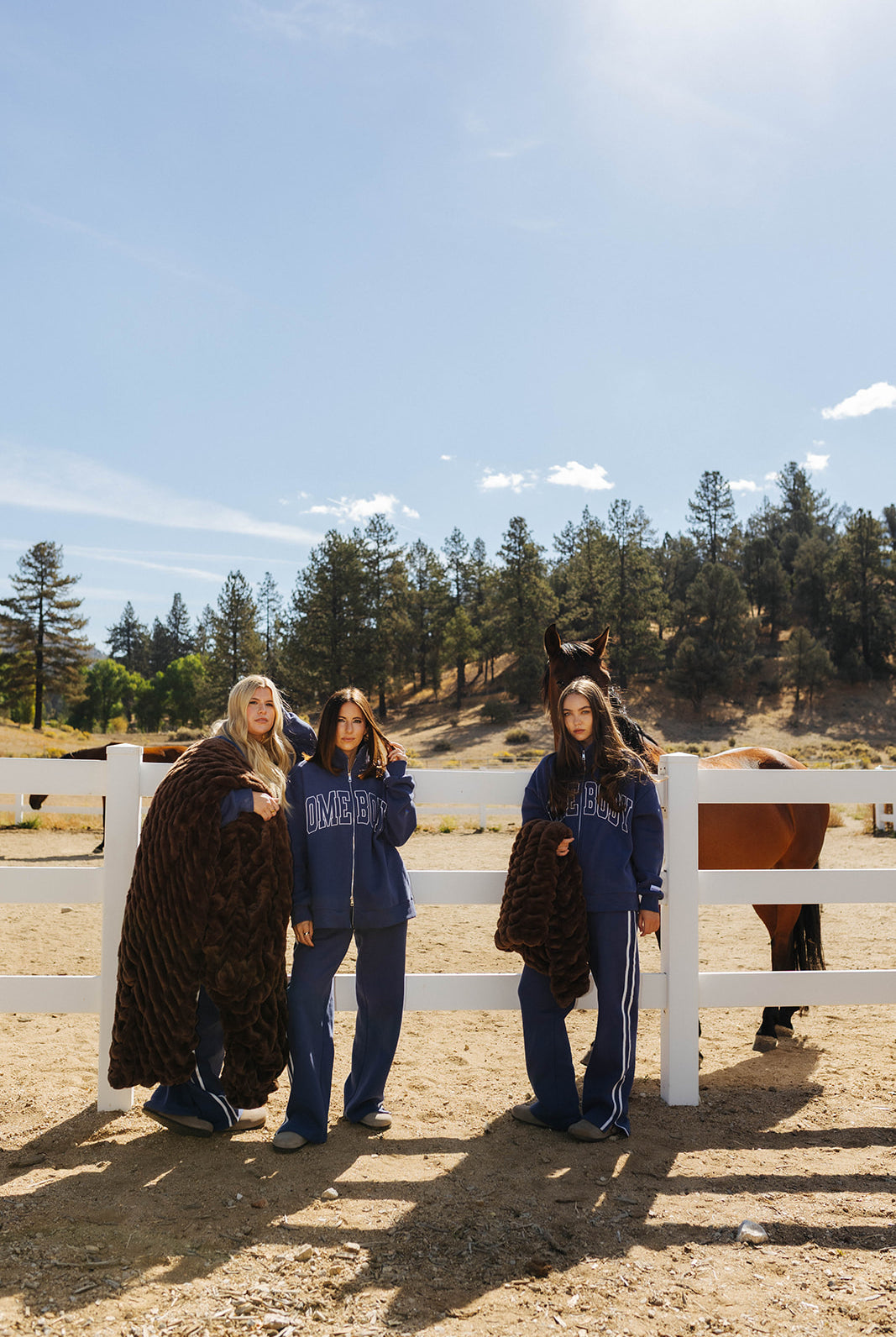 Three models displaying chocolate brown faux fur blankets beside a white fence.