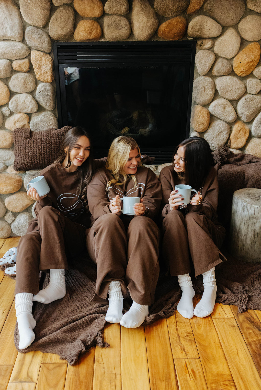 Three models wearing brown sets and sitting together on a brown waffle fringe blanket beside a stone fireplace.