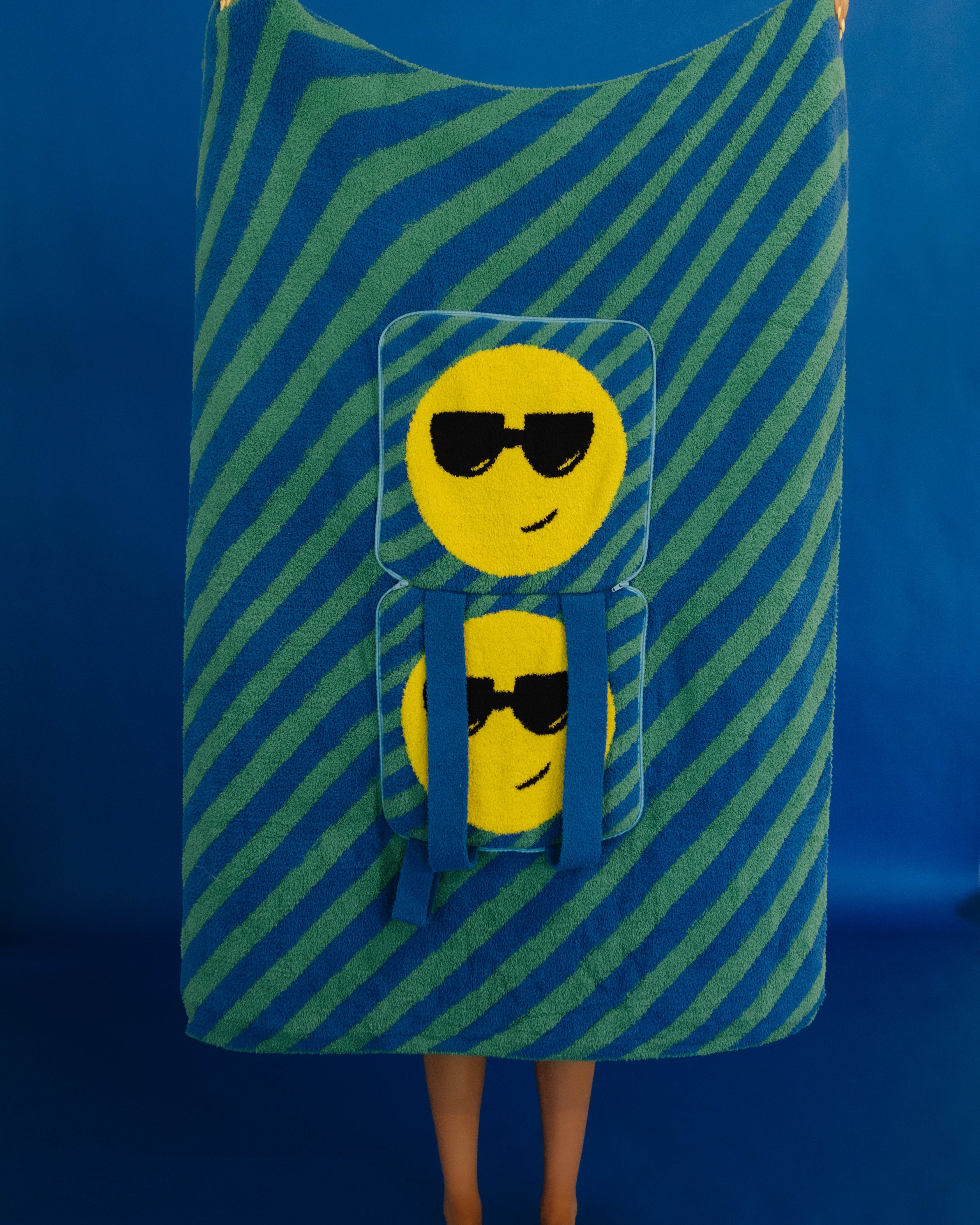 A person standing barefoot against a blue studio backdrop, holding up a large rectangular quillow with diagonal blue and green stripes and two yellow emoji faces wearing sunglasses printed on the front.
