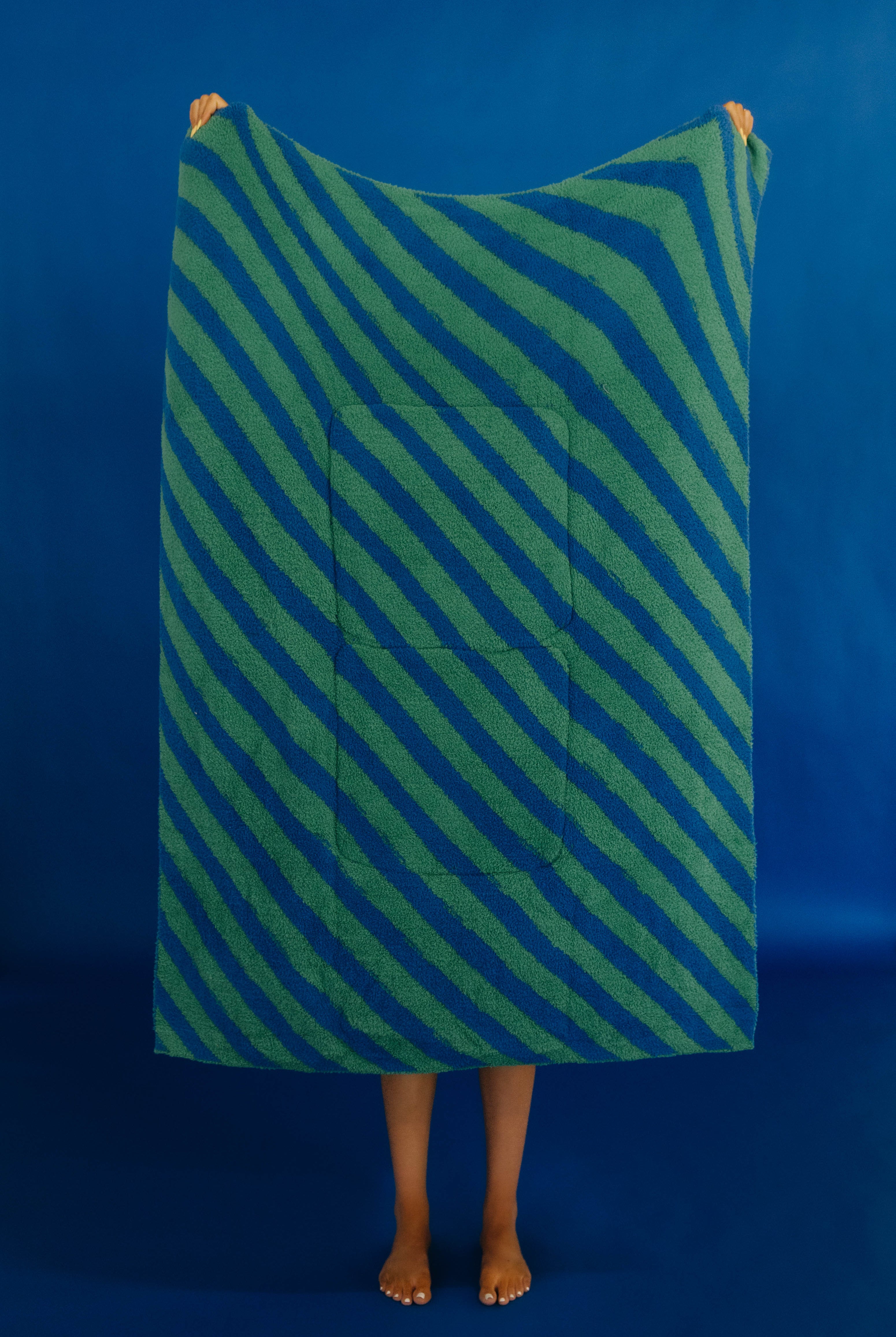 A person standing barefoot against a solid blue backdrop, holding up a large rectangular quillow with diagonal green and blue stripes and a central pocket.