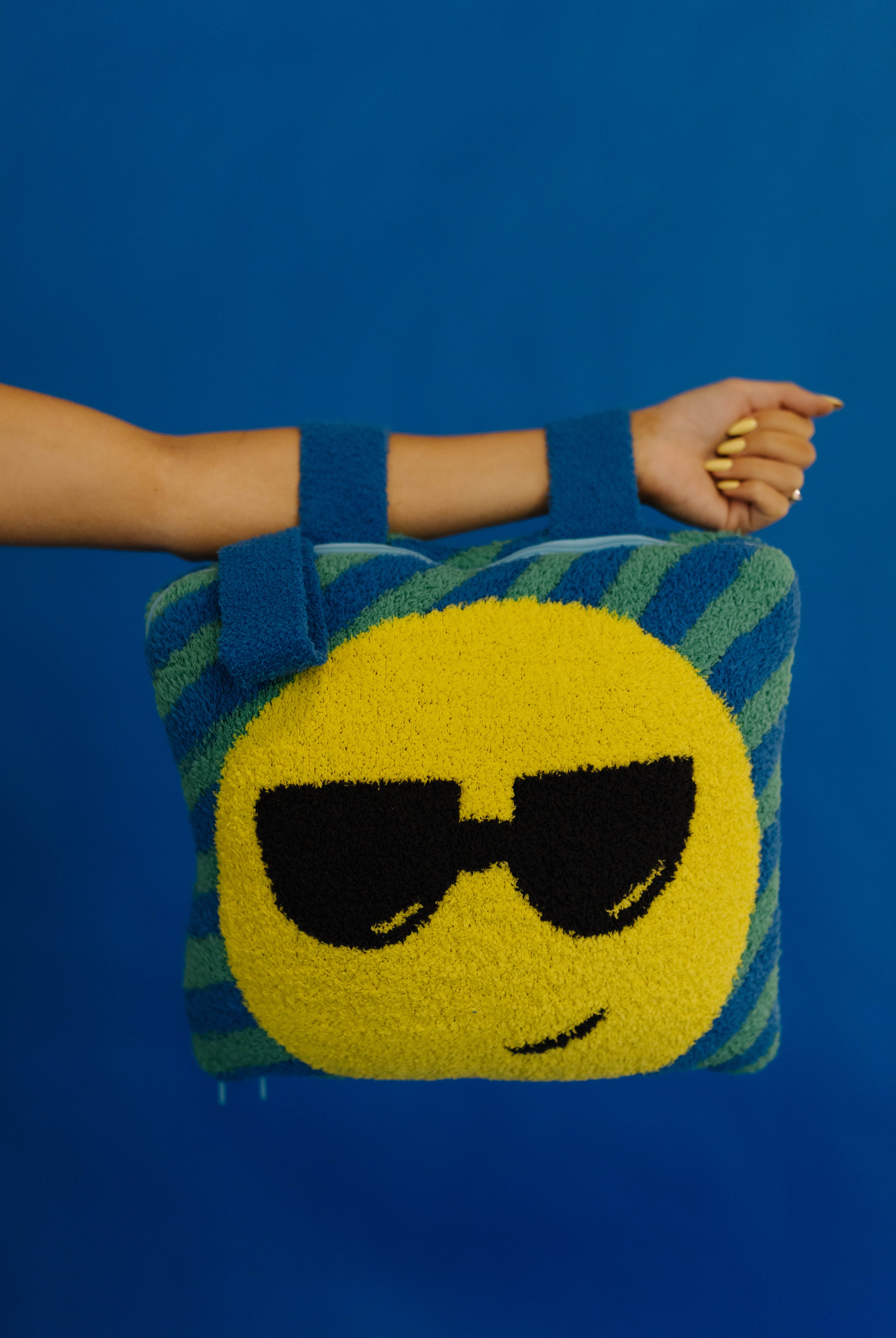 A person's arm and hand holding a quillow with black sunglasses and blue striped sides, set against a solid blue background.