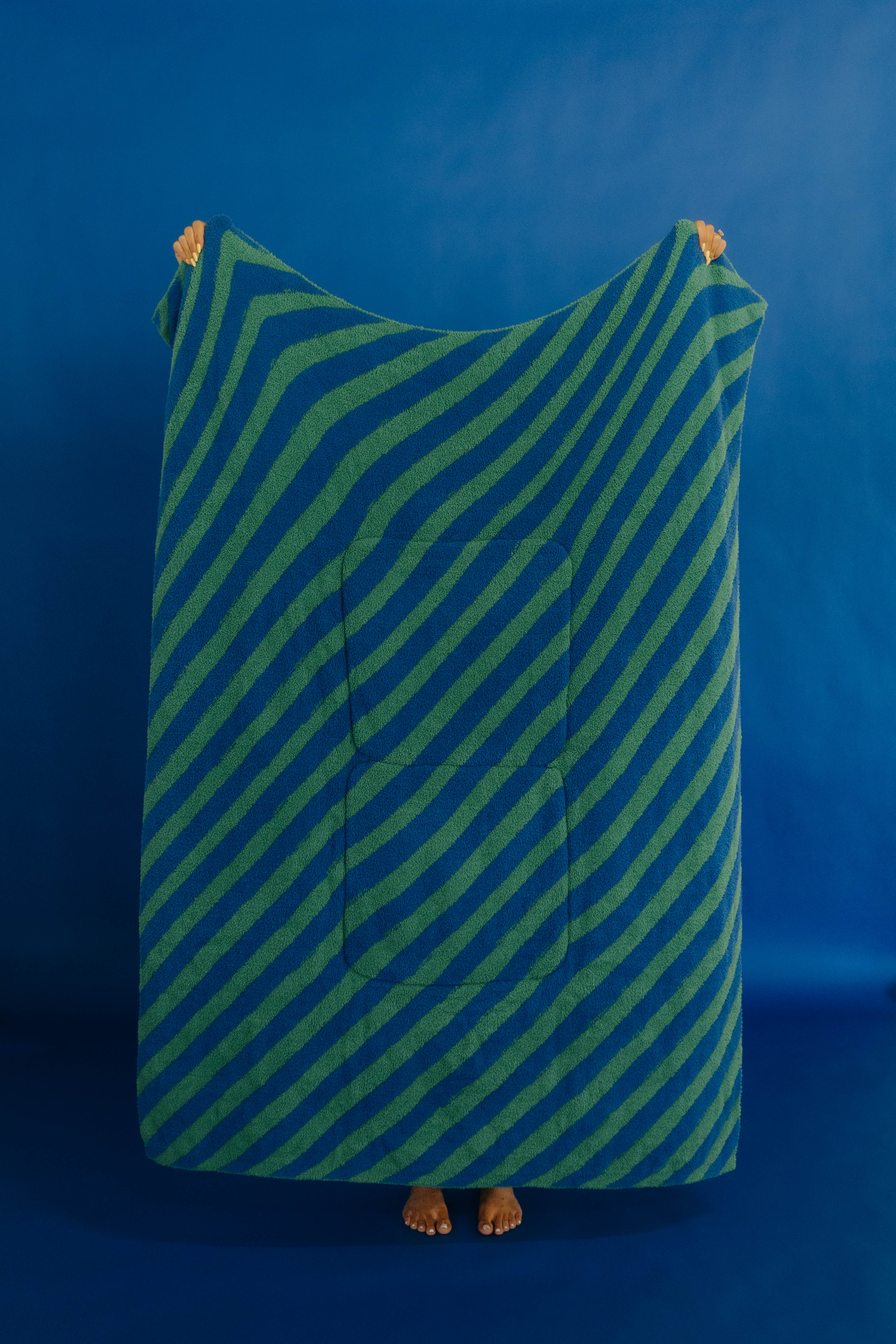 A person standing barefoot against a blue studio background, holding up a large rectangular towel or blanket with diagonal blue and green stripes and a central pocket.