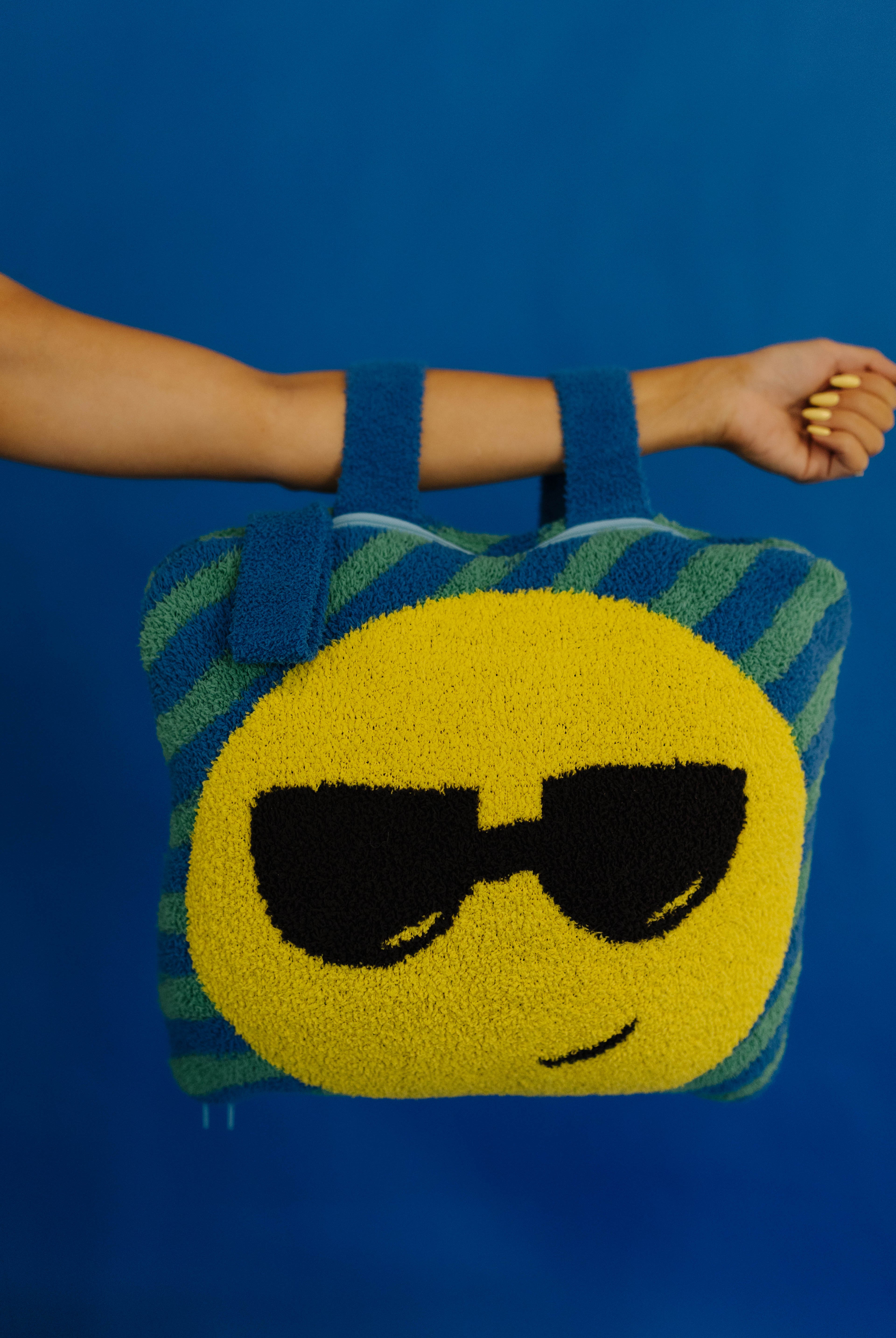 A person's arm and hand a quillow with a yellow smiley face wearing black sunglasses, set against a solid blue background.