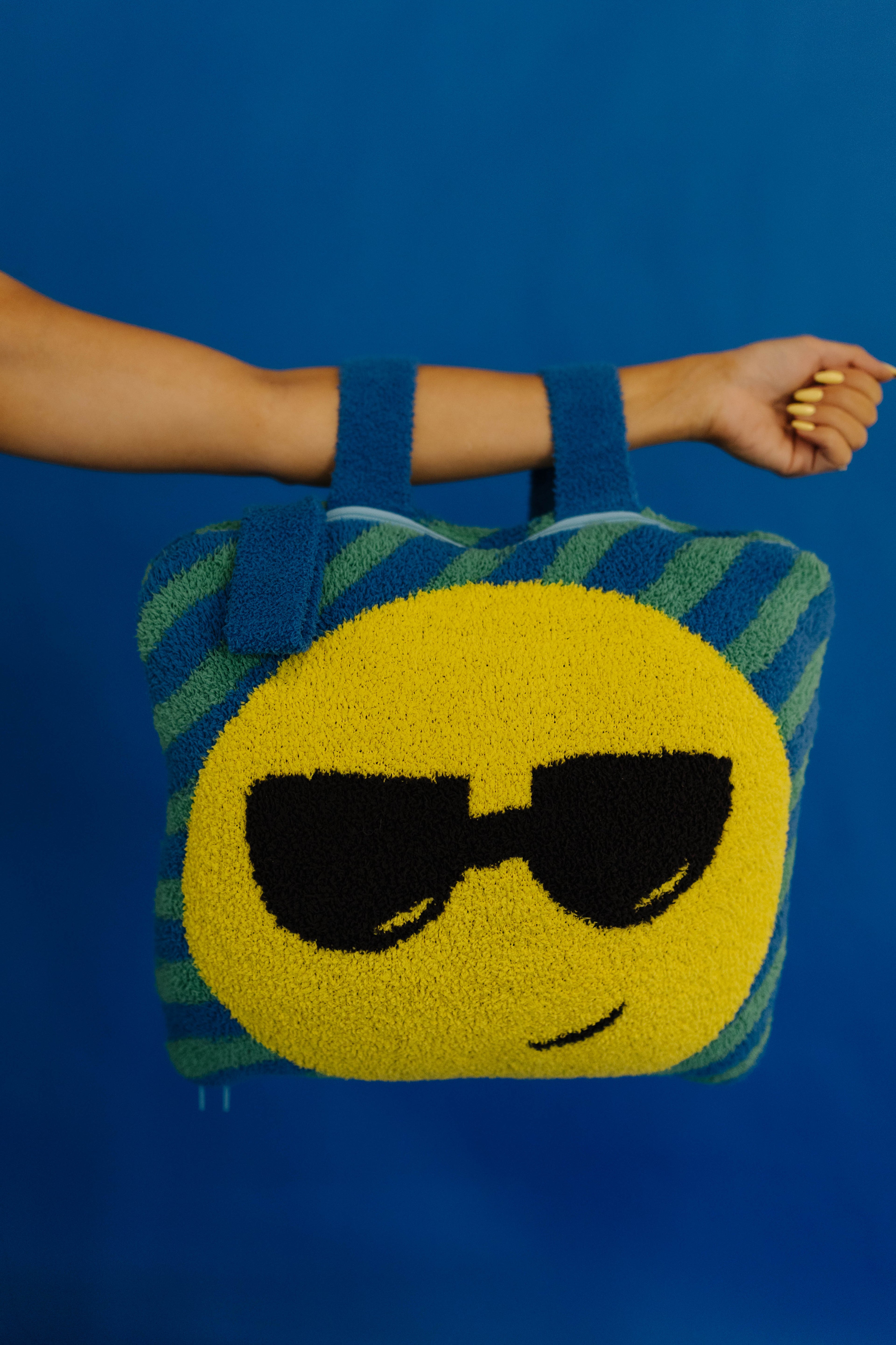 A person's arm and hand a quillow with a yellow smiley face wearing black sunglasses, set against a solid blue background.
