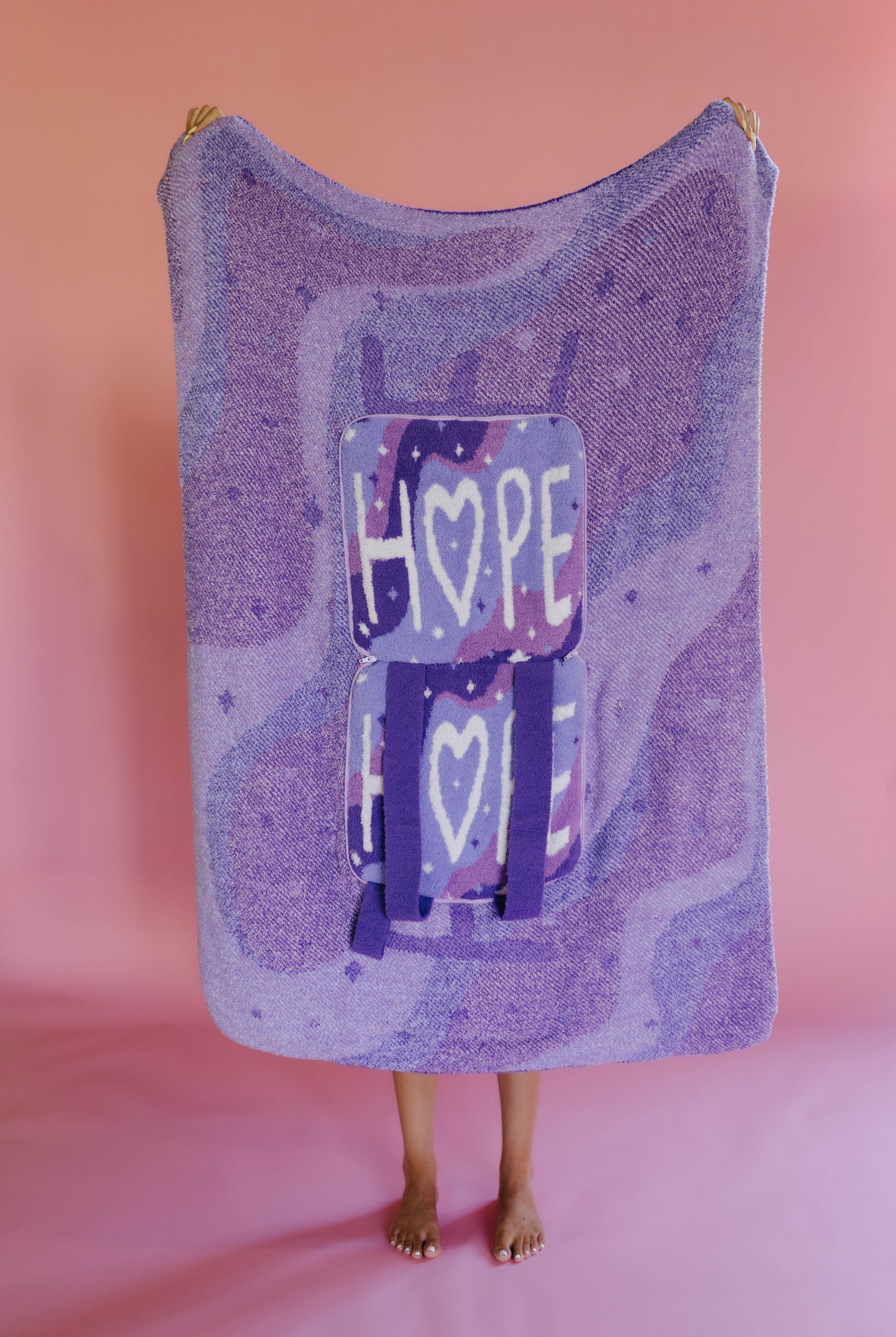 A person holding up a quillow, with backpack straps and a graphic featuring the word 'HOPE' and hearts, standing against a solid pink backdrop.