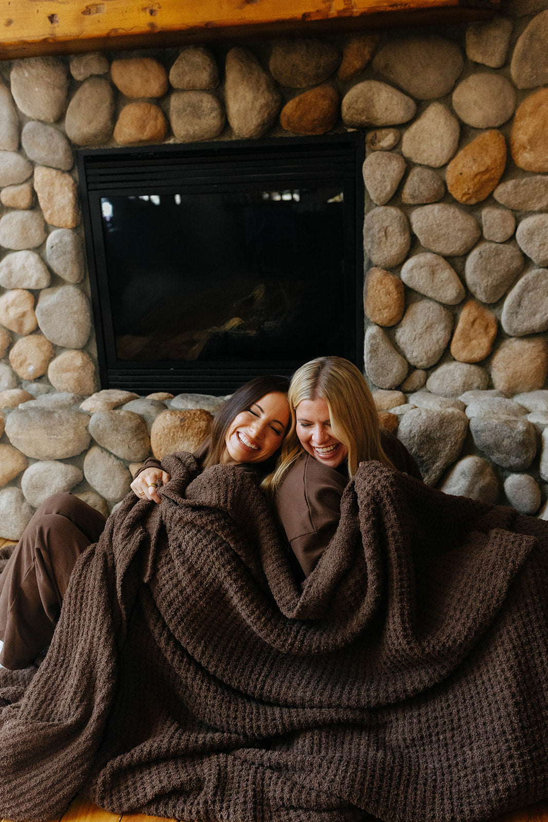 Two models sharing the brown waffle blanket while seated by a fireplace.