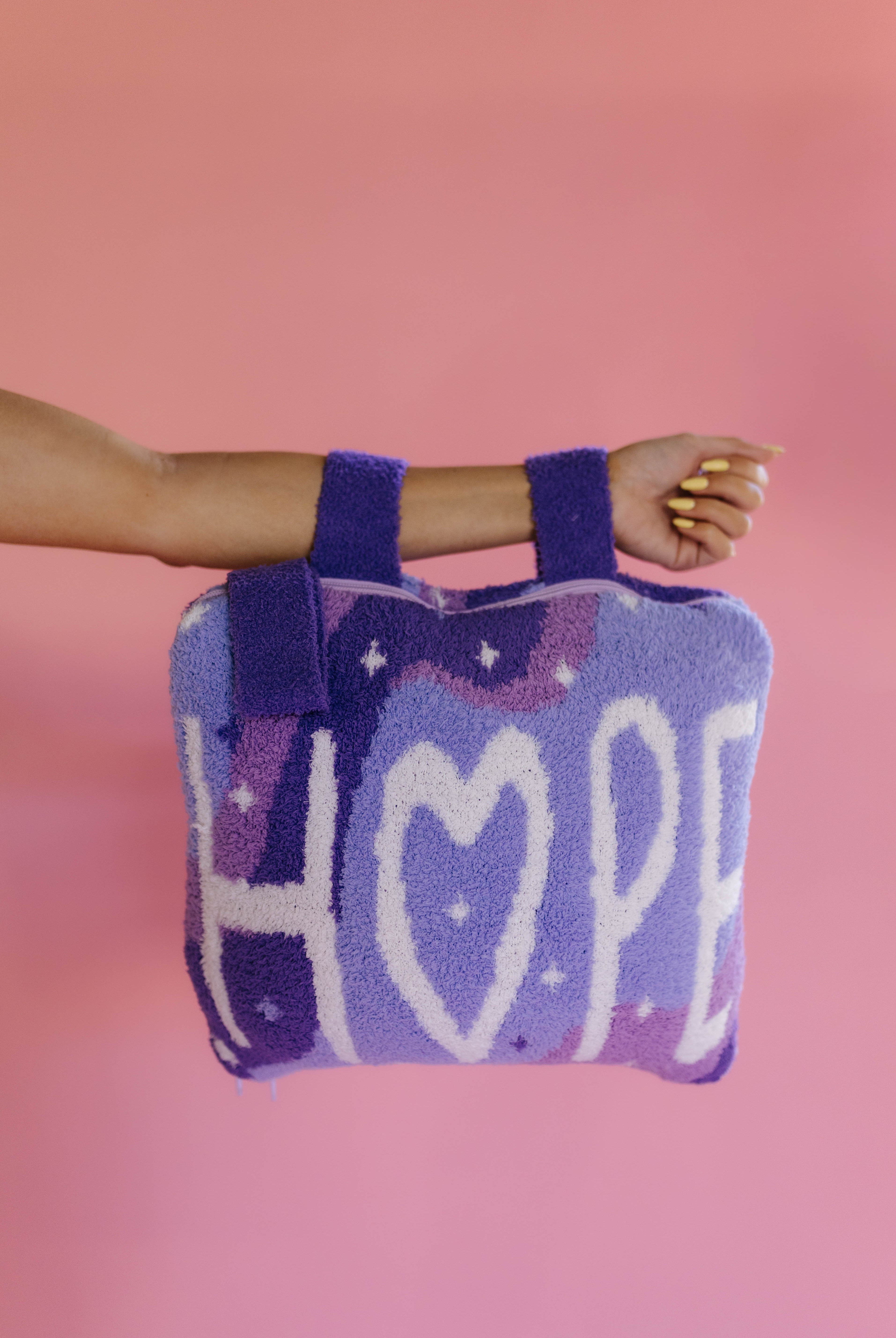 A person's arm and hand holding a purple and lavender quillow with the word 'HOPE' knitted in white, set against a solid pink background.
