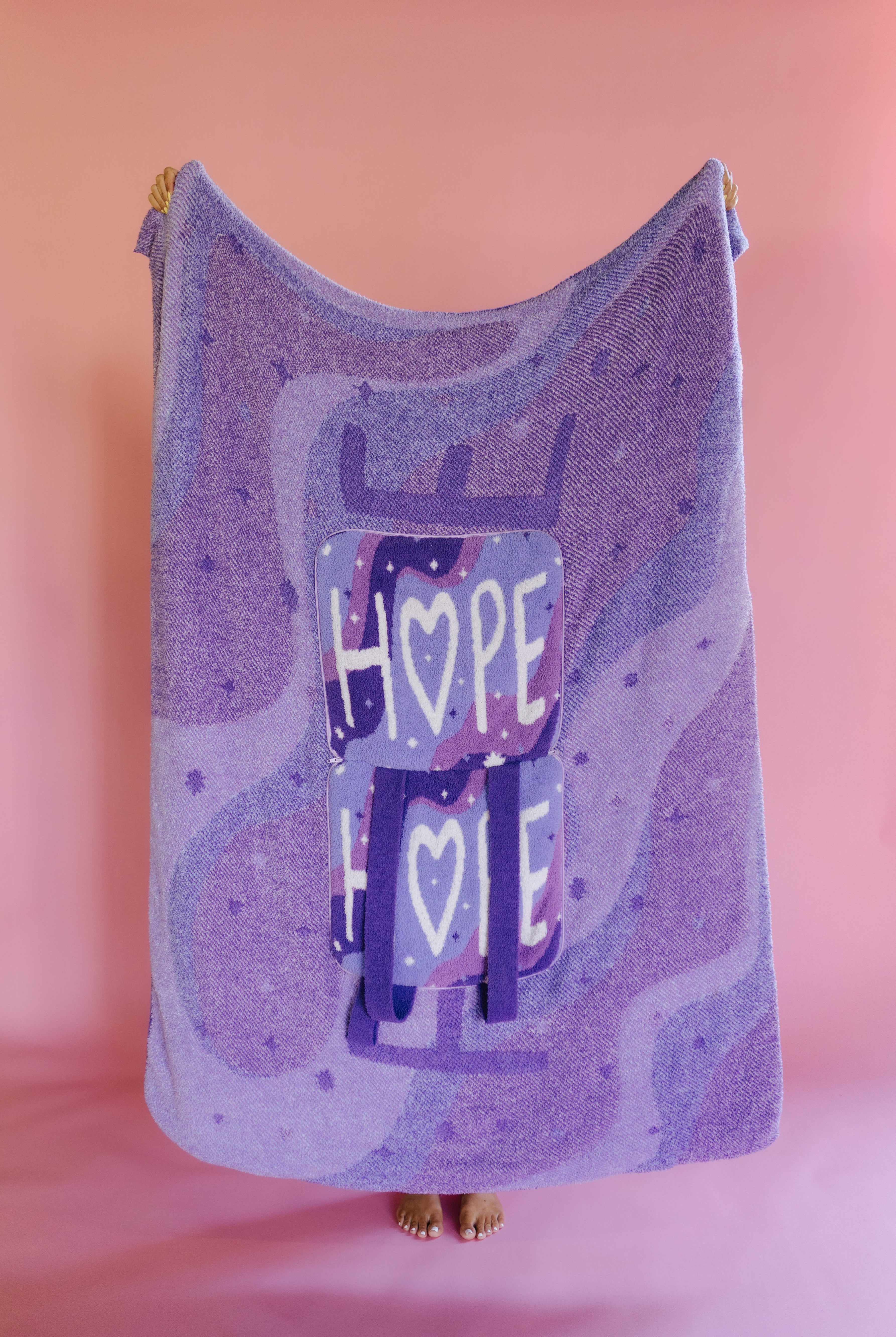 A lavender and purple quillow with a central graphic of stacked boxes spelling 'HOPE' with hearts, held up against a solid pink background.
