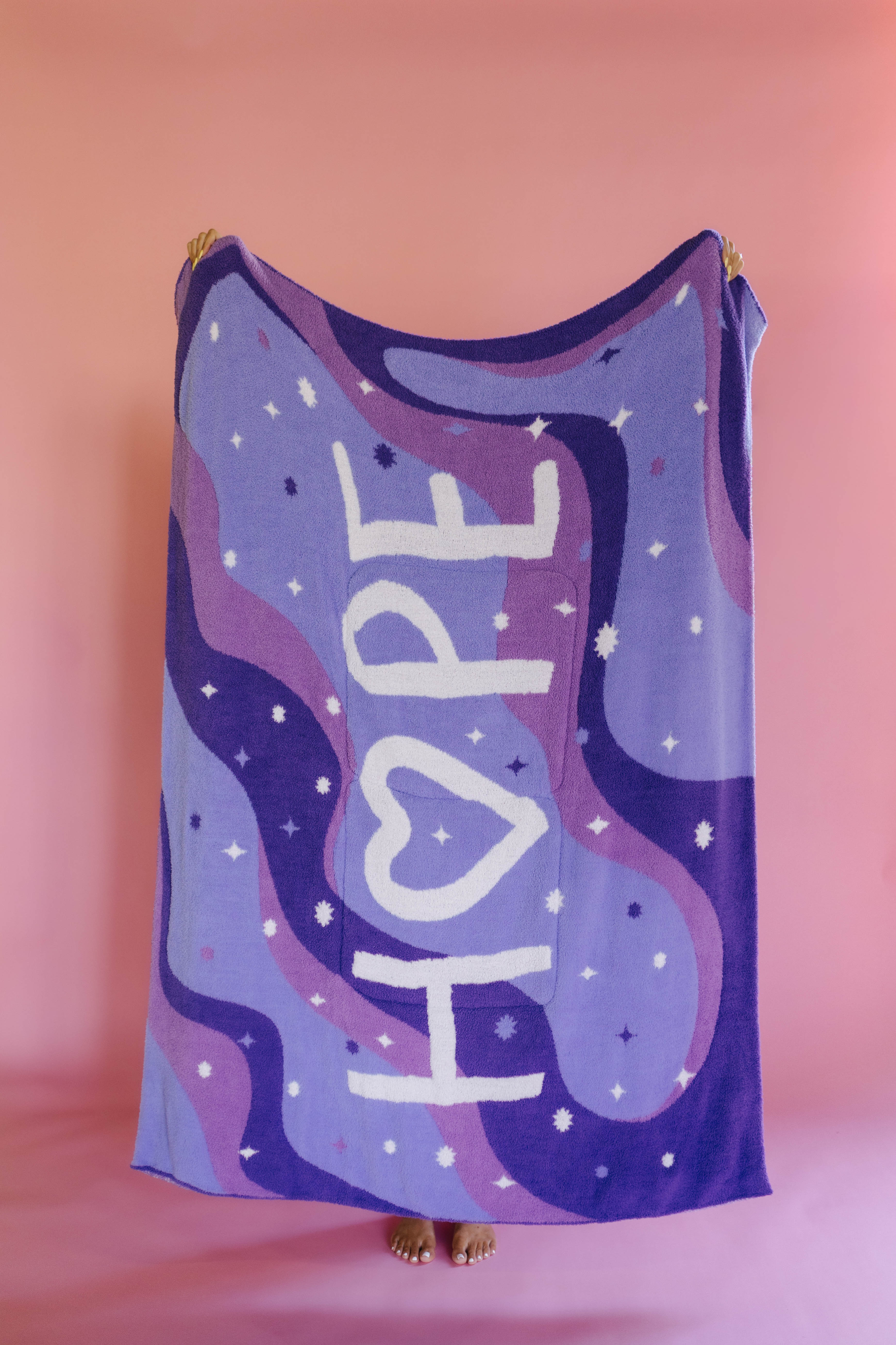 A person holding up the full size quillow with white stars, and the word 'HOPE' in large white letters, standing against a pink background.