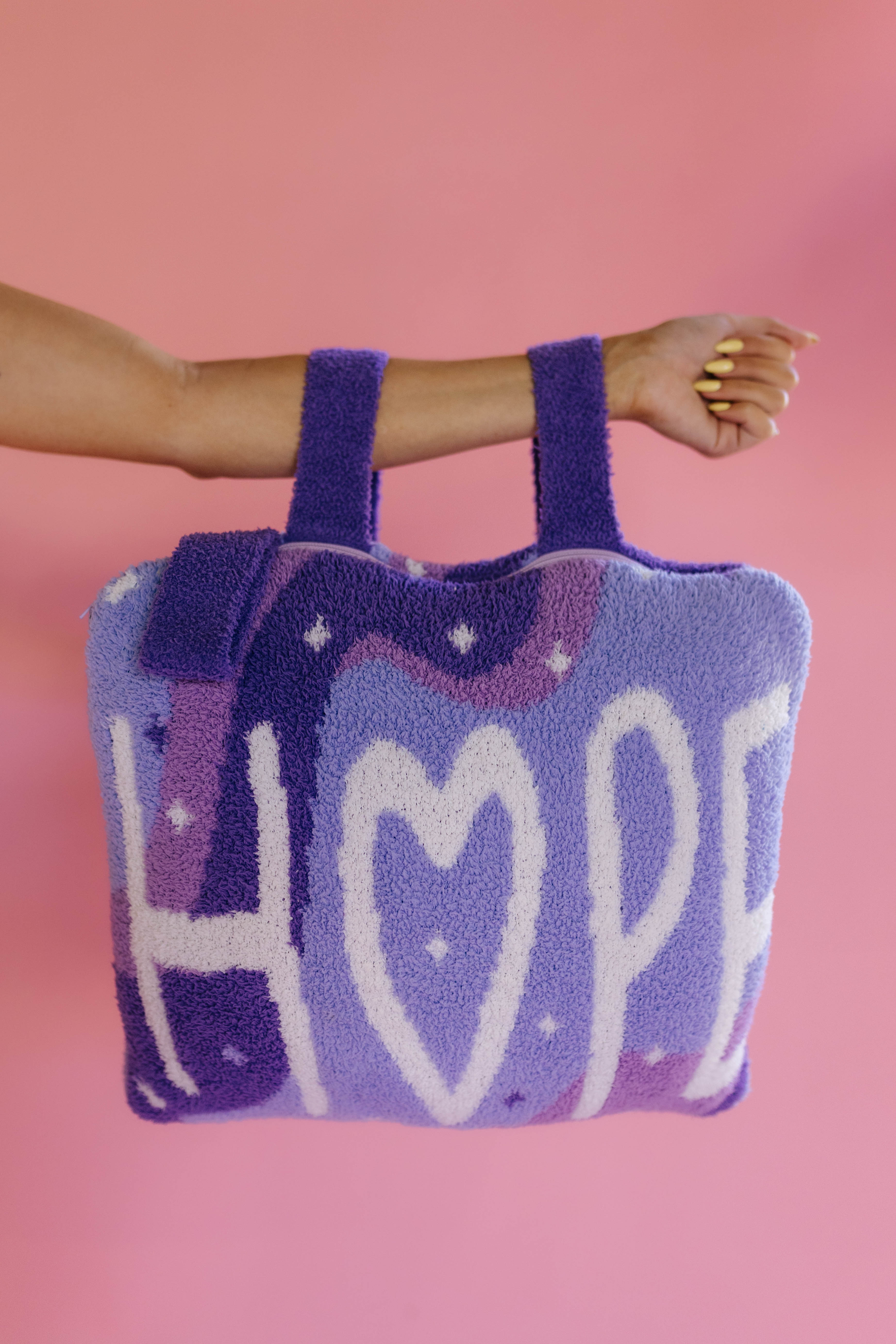 A fuzzy lavender quillow with the word 'HOPE' in white letters, held by a person's arm against a solid pink background.