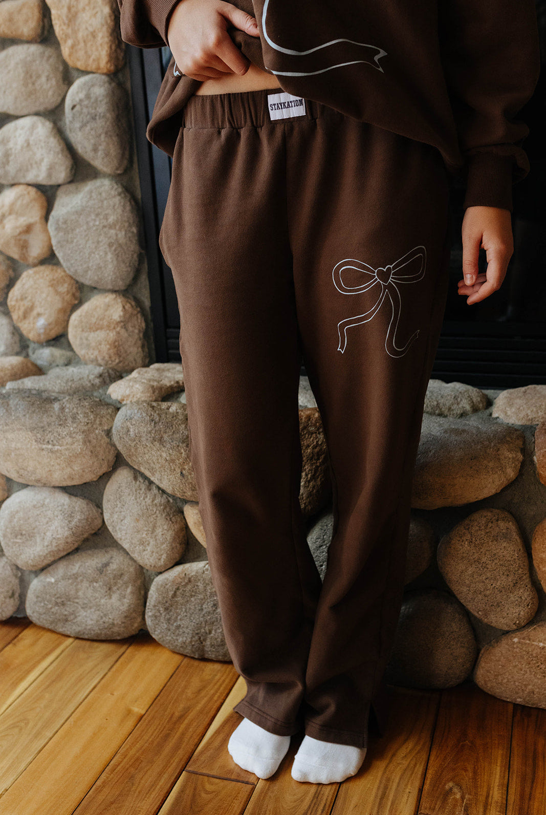Model wearing brown bow sweatpants by a fireplace.