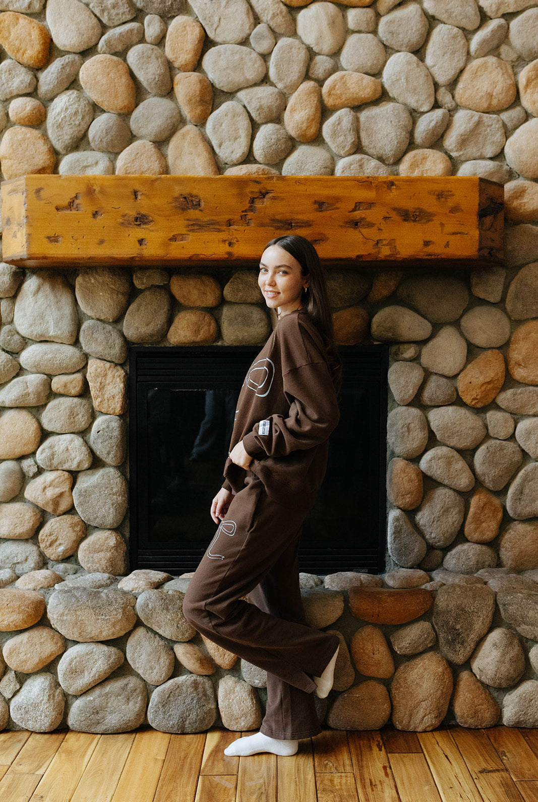 Side and back view of the brown bow sweatshirt and sweatpants by a fireplace.