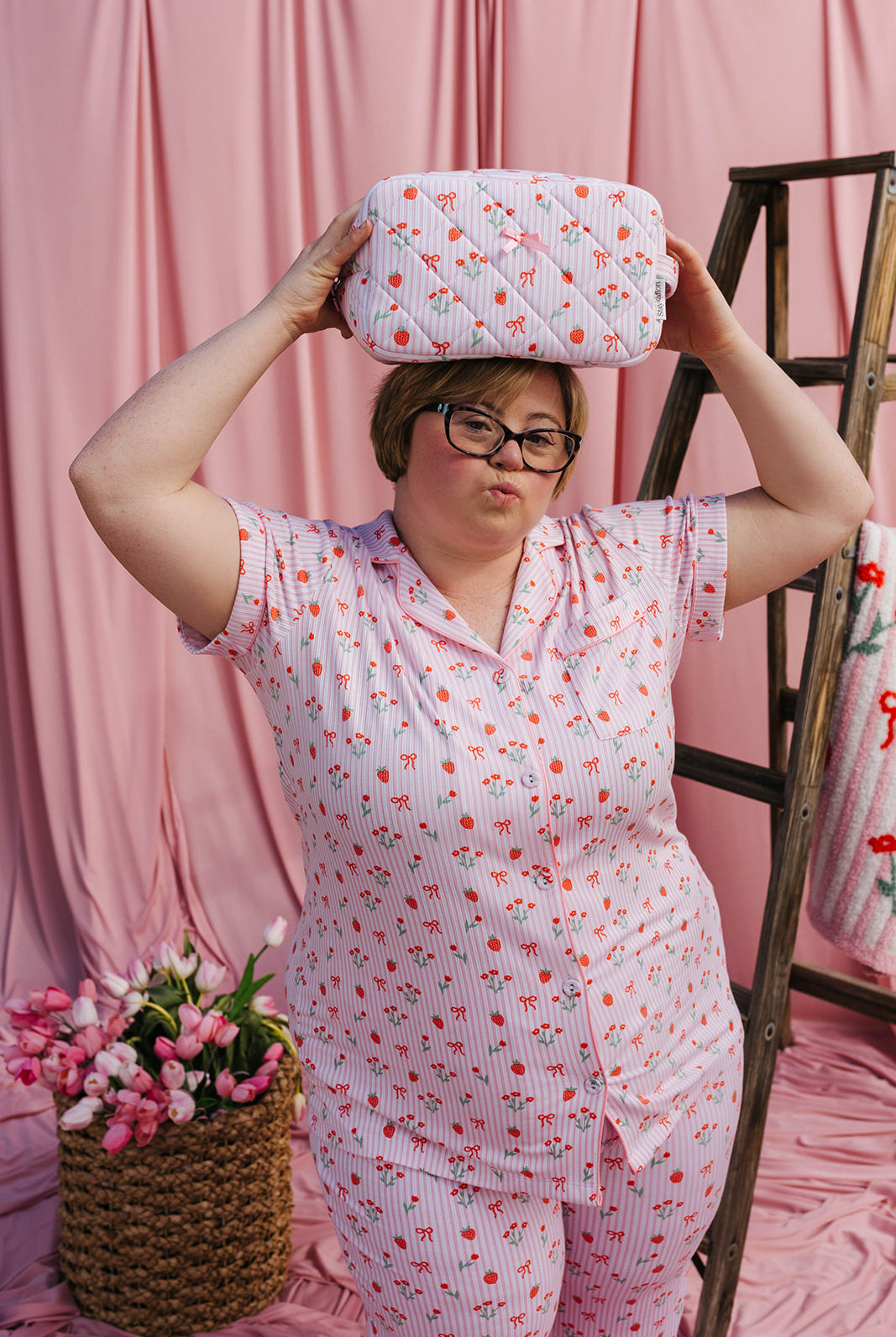 Person wearing a pink pajama set holding a patterned cosmetic bag against a pink curtain background.