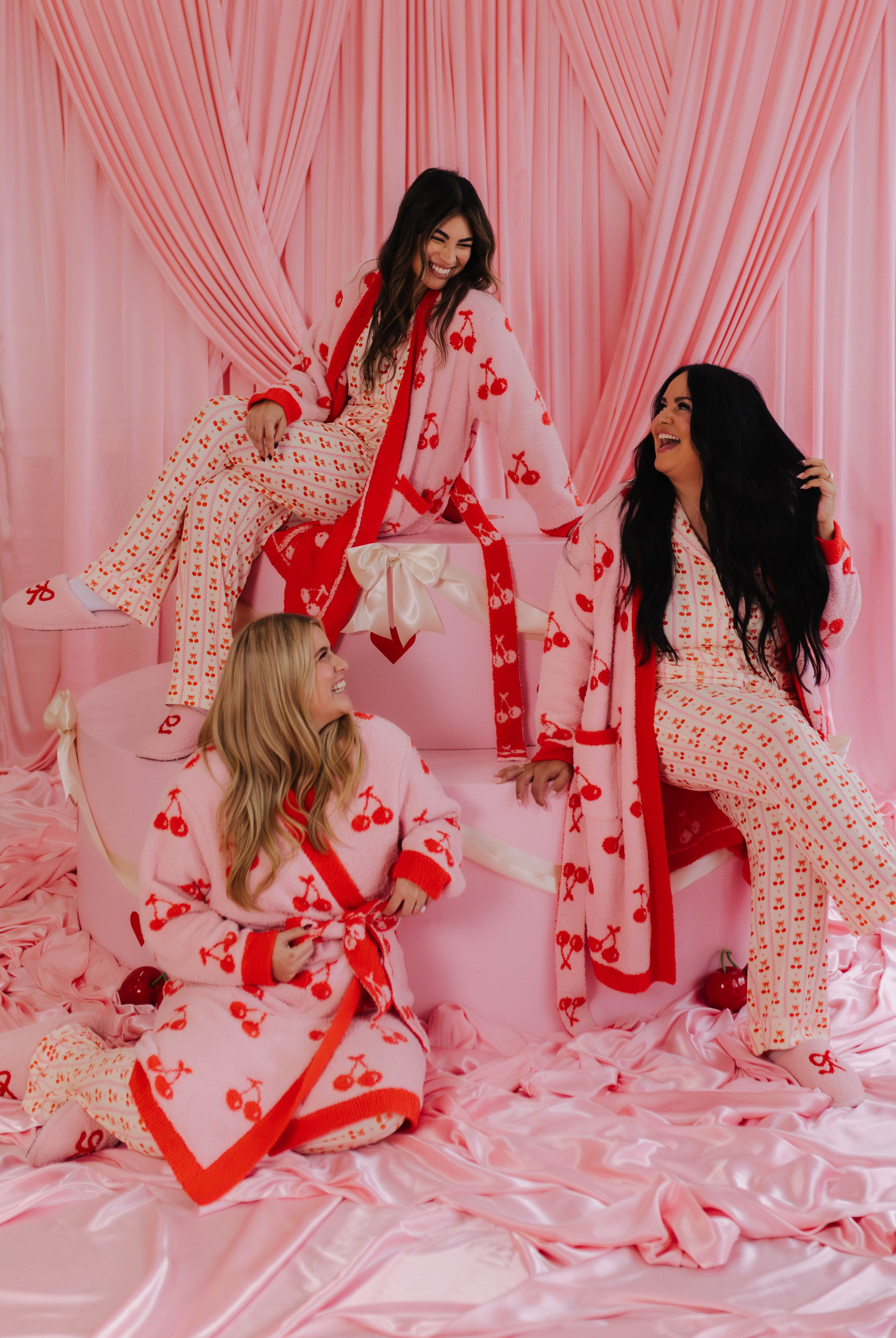 Three women in matching cherry-themed pajamas with a cherry robe sitting on a pink couch with pink curtains in the background.