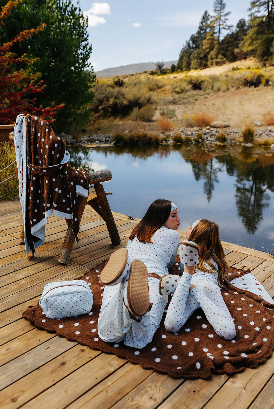 Models in blue polka dot pajamas sitting on a brown polka dot blanket, with folded quillows on a lakeside dock.