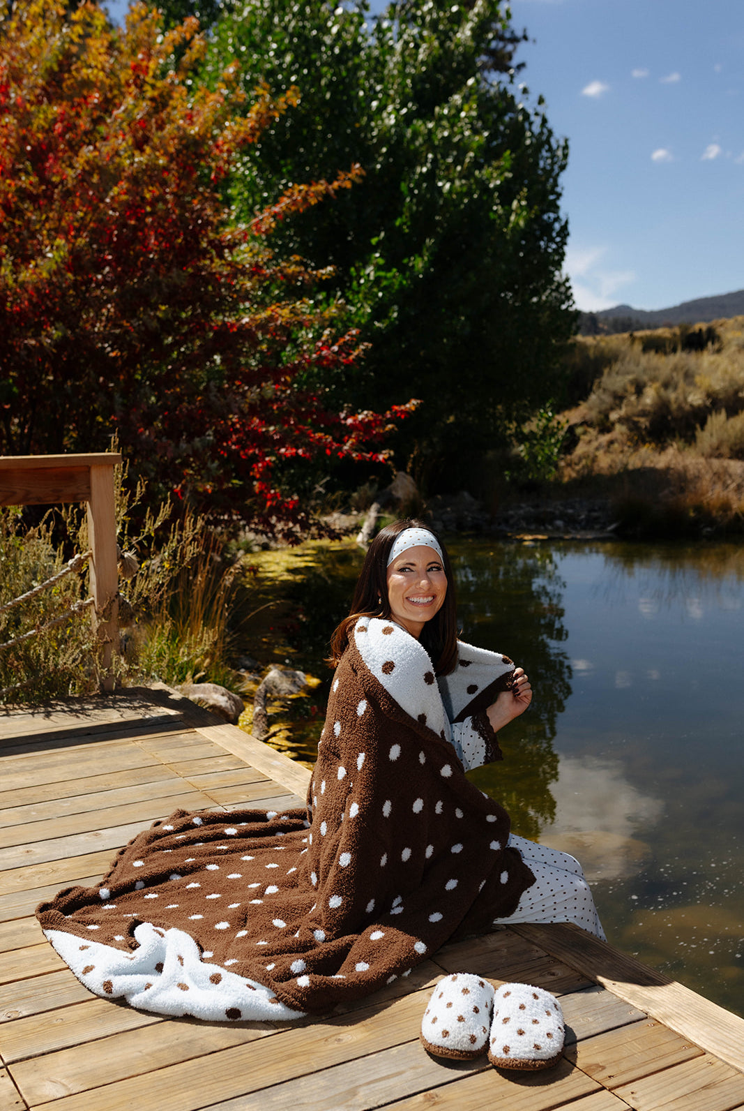 Model seated on a dock wrapped in a brown polka dot quillow blanket, showing its cream reverse with brown dots.