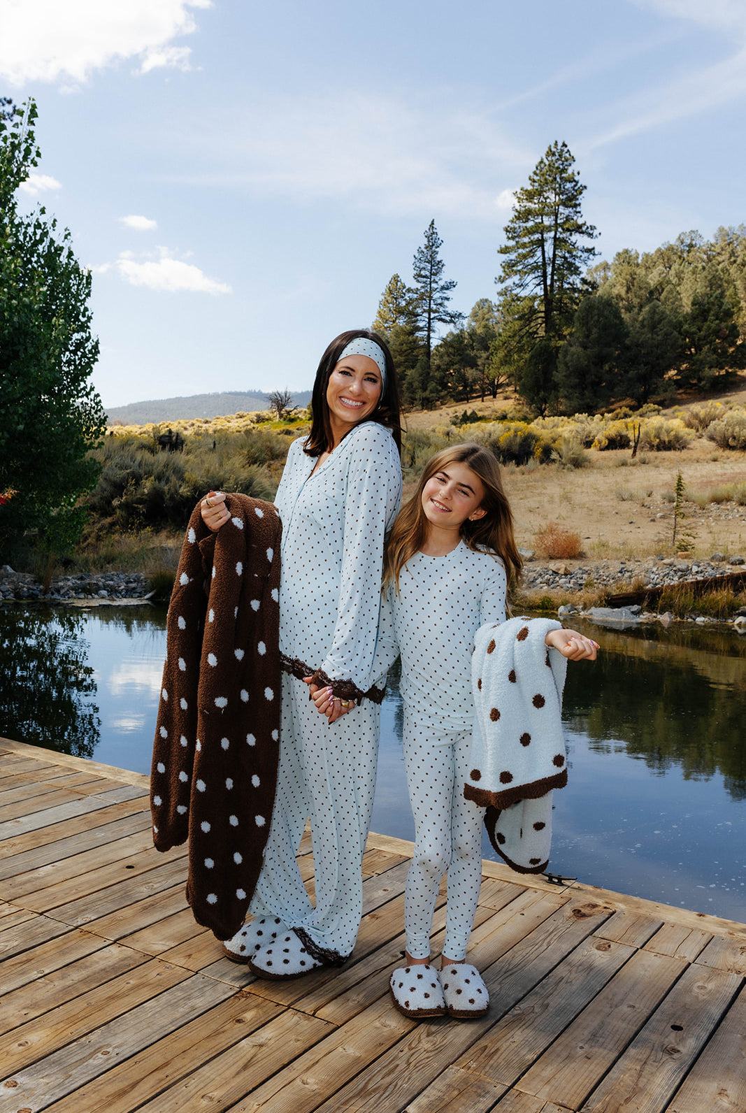 Adult and child in blue polka dot pajamas holding brown polka dot blankets with cream reverse sides beside a lake.