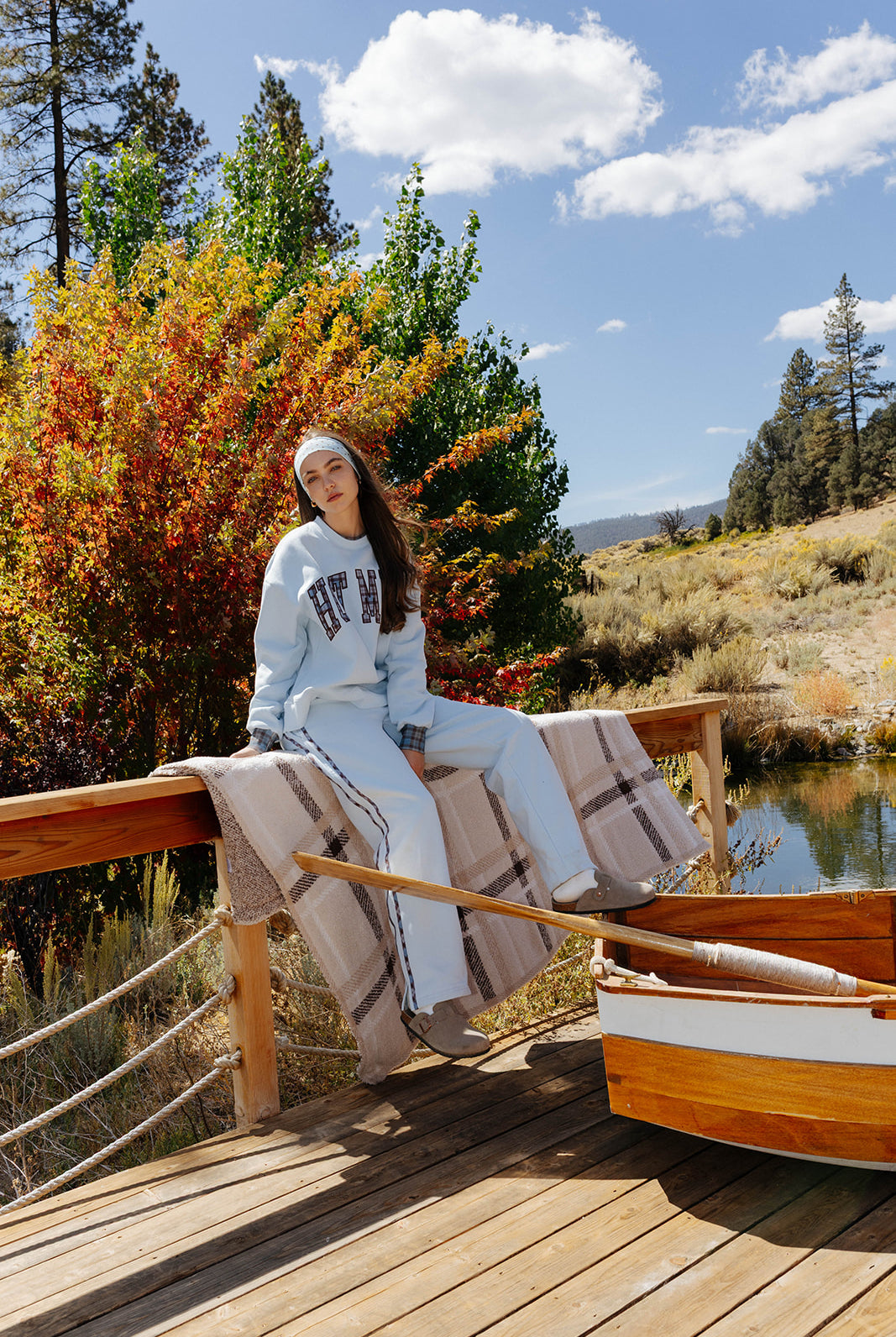 Model in a light blue Hot Mess set sitting on a wooden boat with a beige and brown plaid blanket beside a lake.