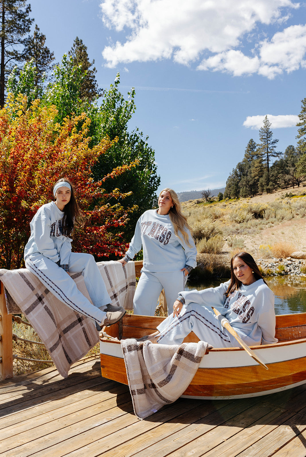 Three models in light blue Hot Mess sweatshirts and sweatpants beside a boat and plaid blanket.