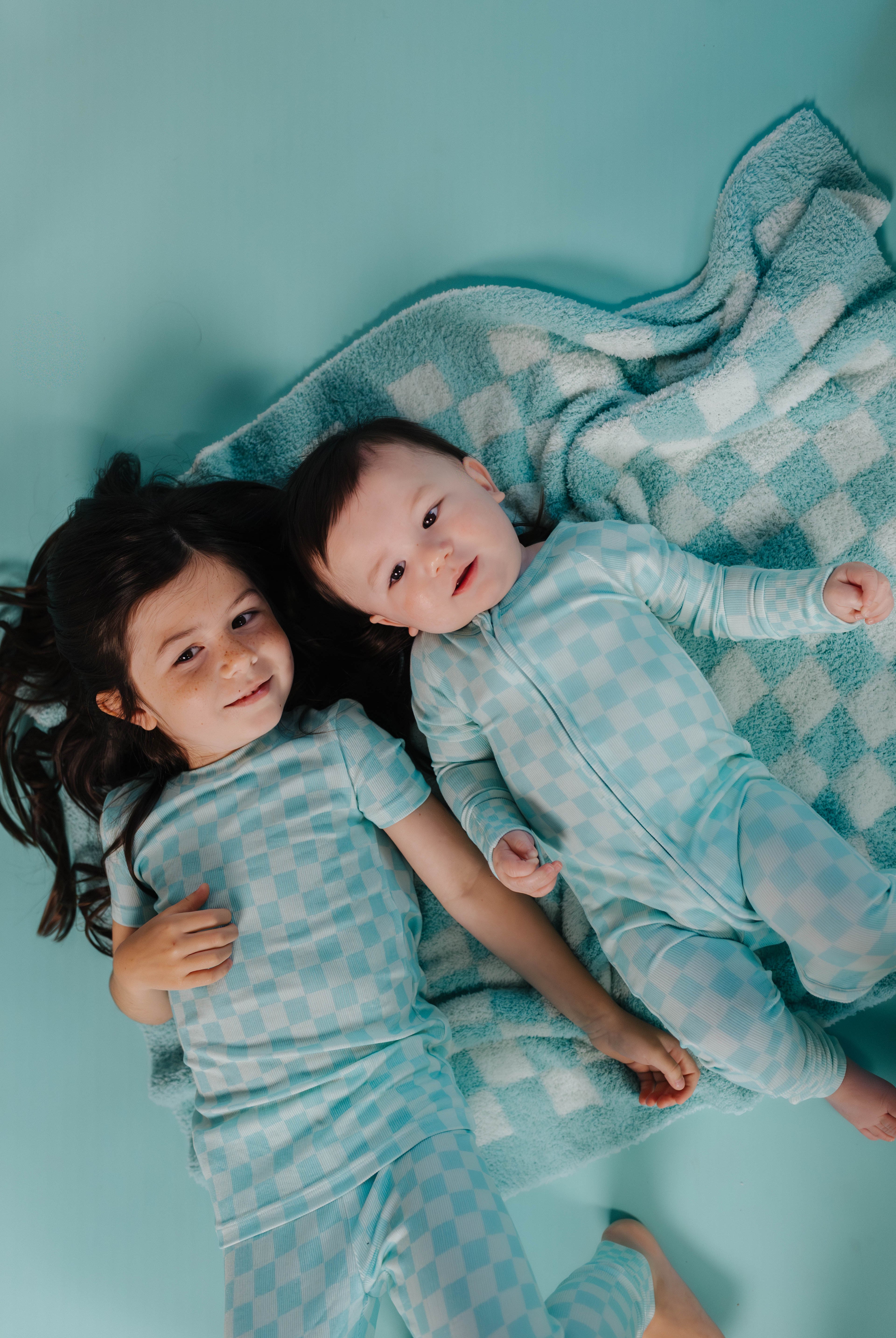 Two children in matching pajamas lying on a checkered blanket against a teal background