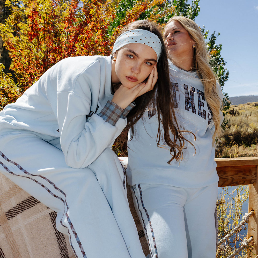 Two models wearing light blue Hot Mess sets with a beige and brown plaid blanket beside a lake.