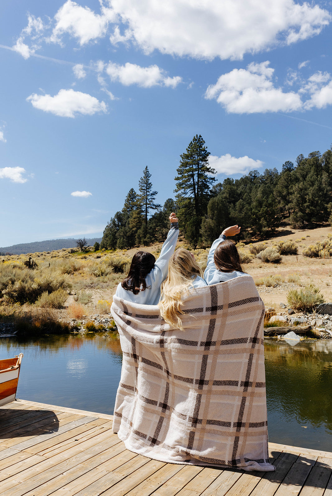 Rear view of three models wrapped together in a beige and brown plaid blanket on a lakeside dock.