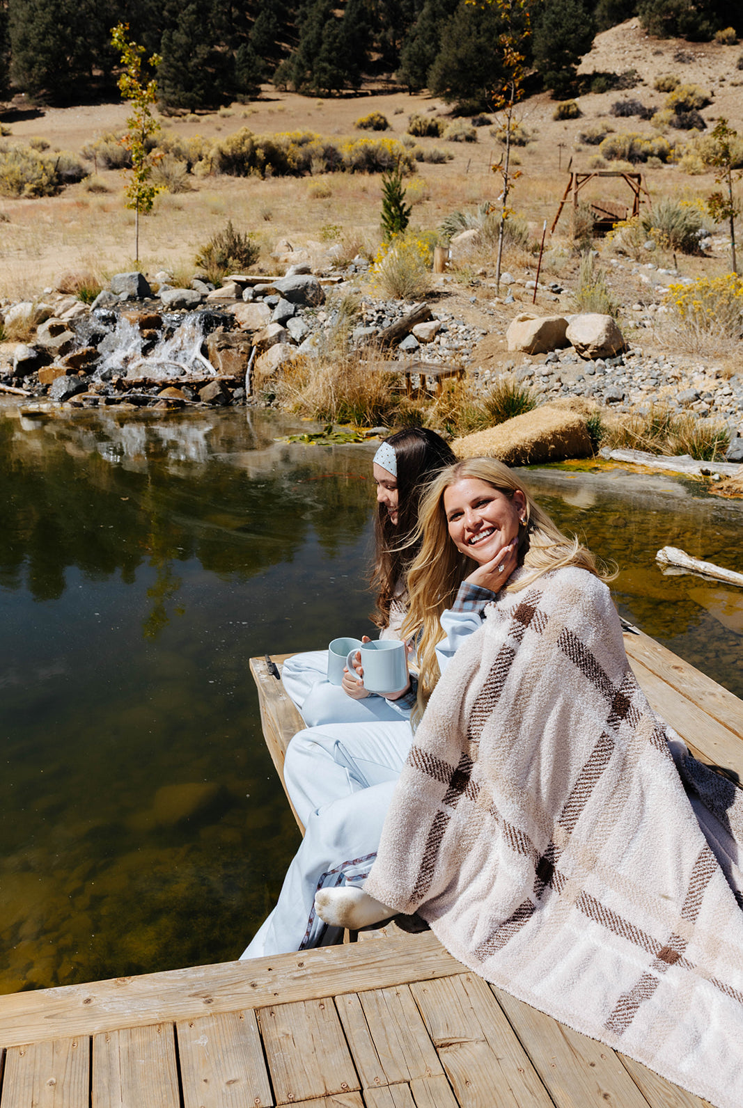 Two models sitting beside a lake with a beige and brown plaid blanket draped around their shoulders.