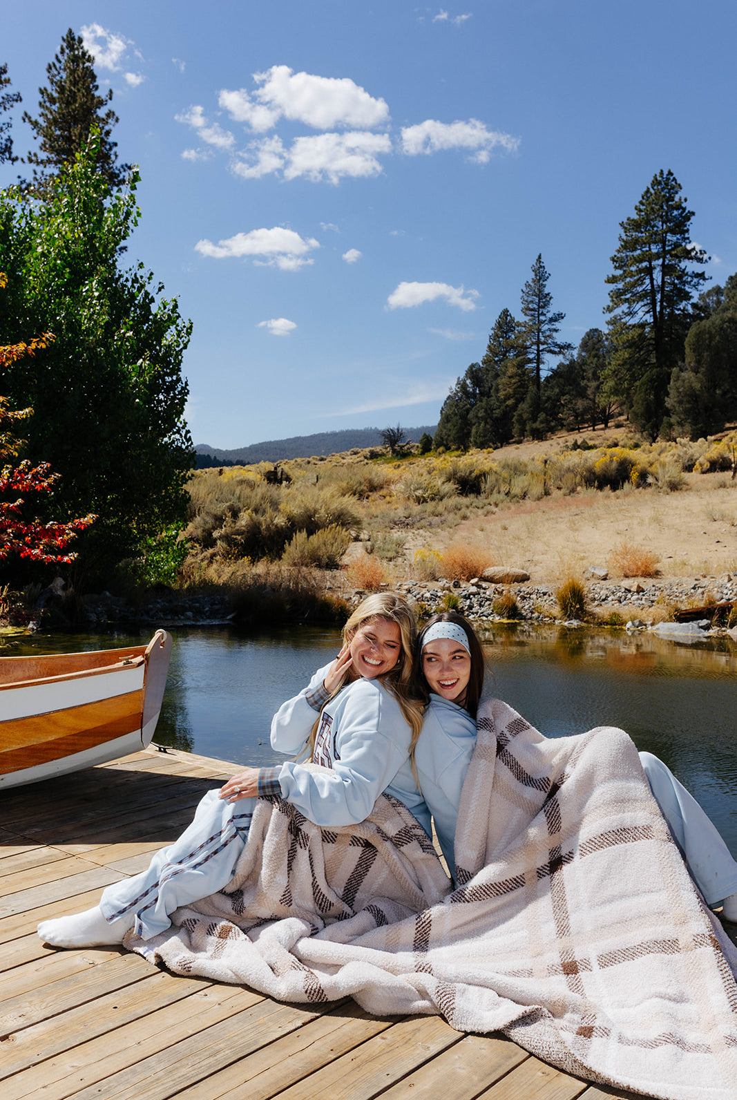 Three models seated together on a dock with the plaid blanket across their laps.