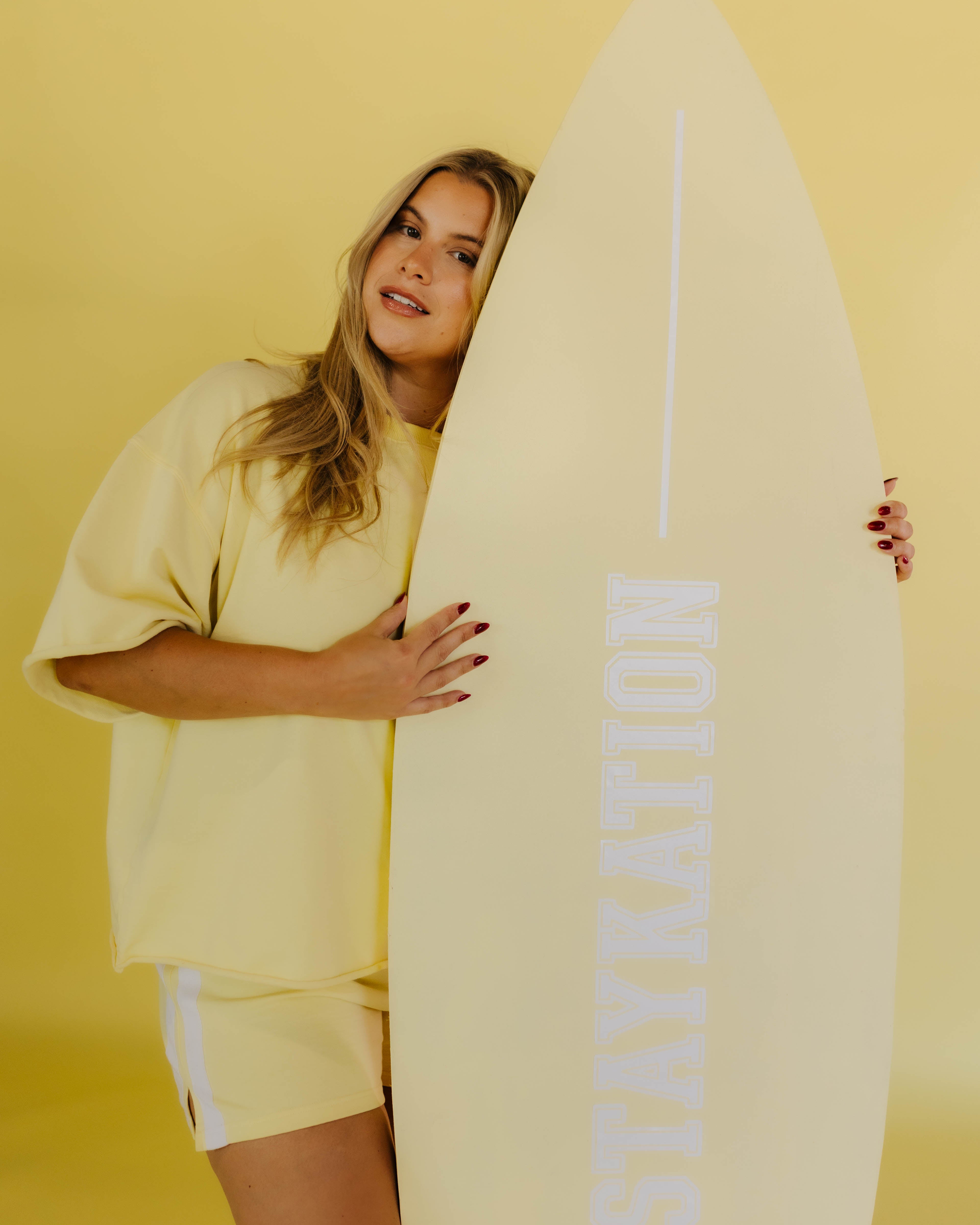 Woman holding a surfboard with 'STAYKATION' branding against a yellow background