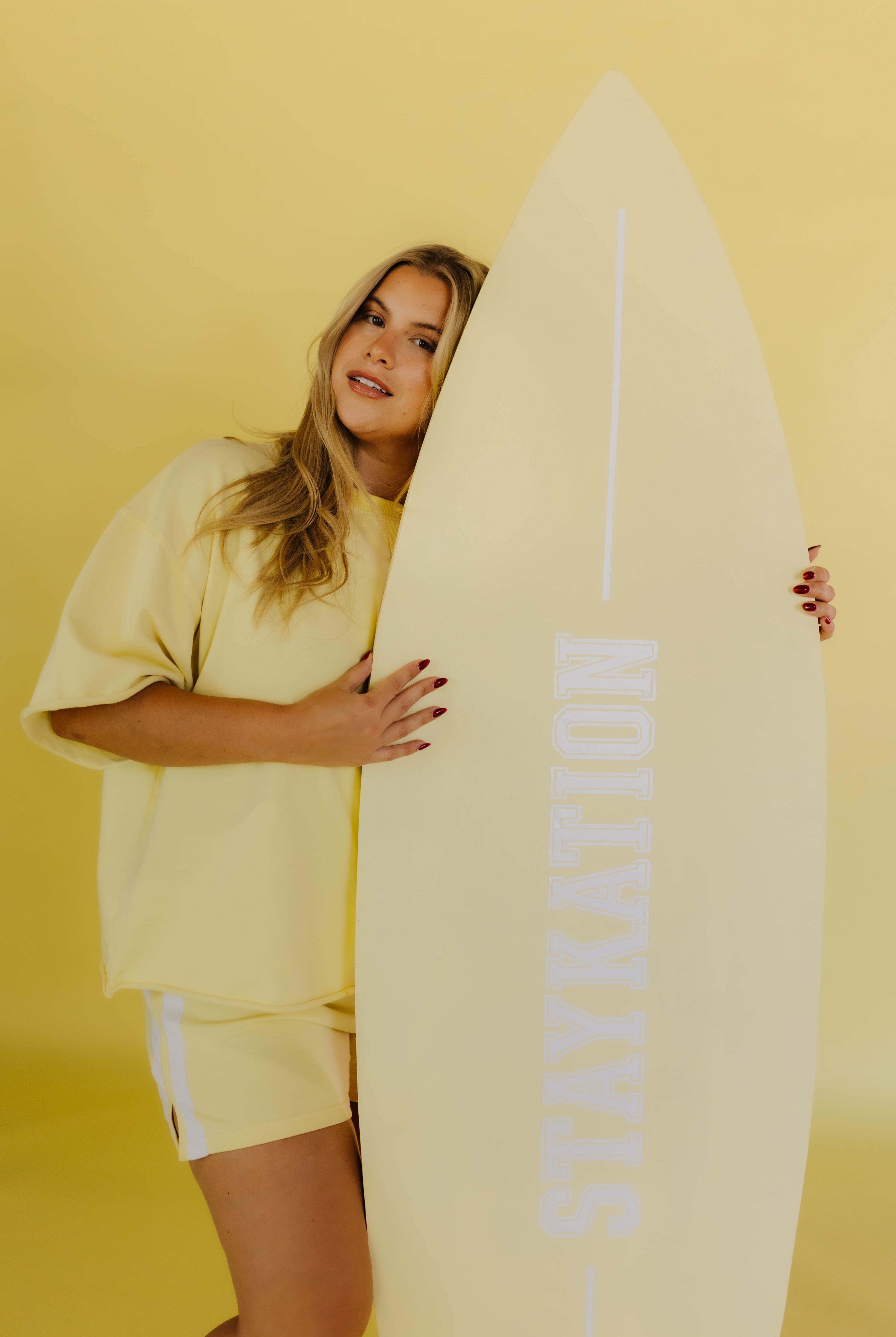 Woman holding a surfboard with 'STAYKATION' branding against a yellow background