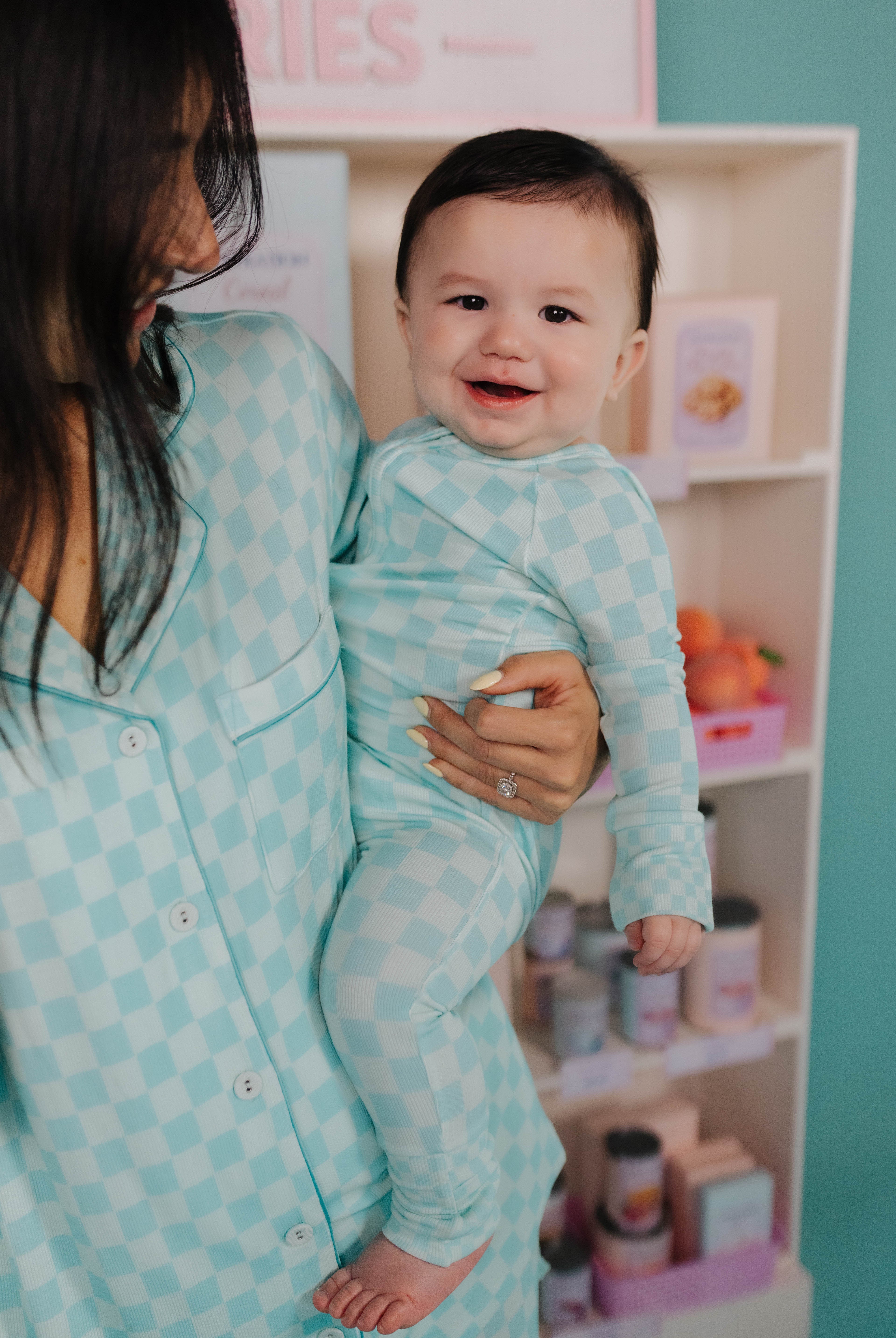 Woman holding a baby in a teal check outfit in a room with shelves in the background.