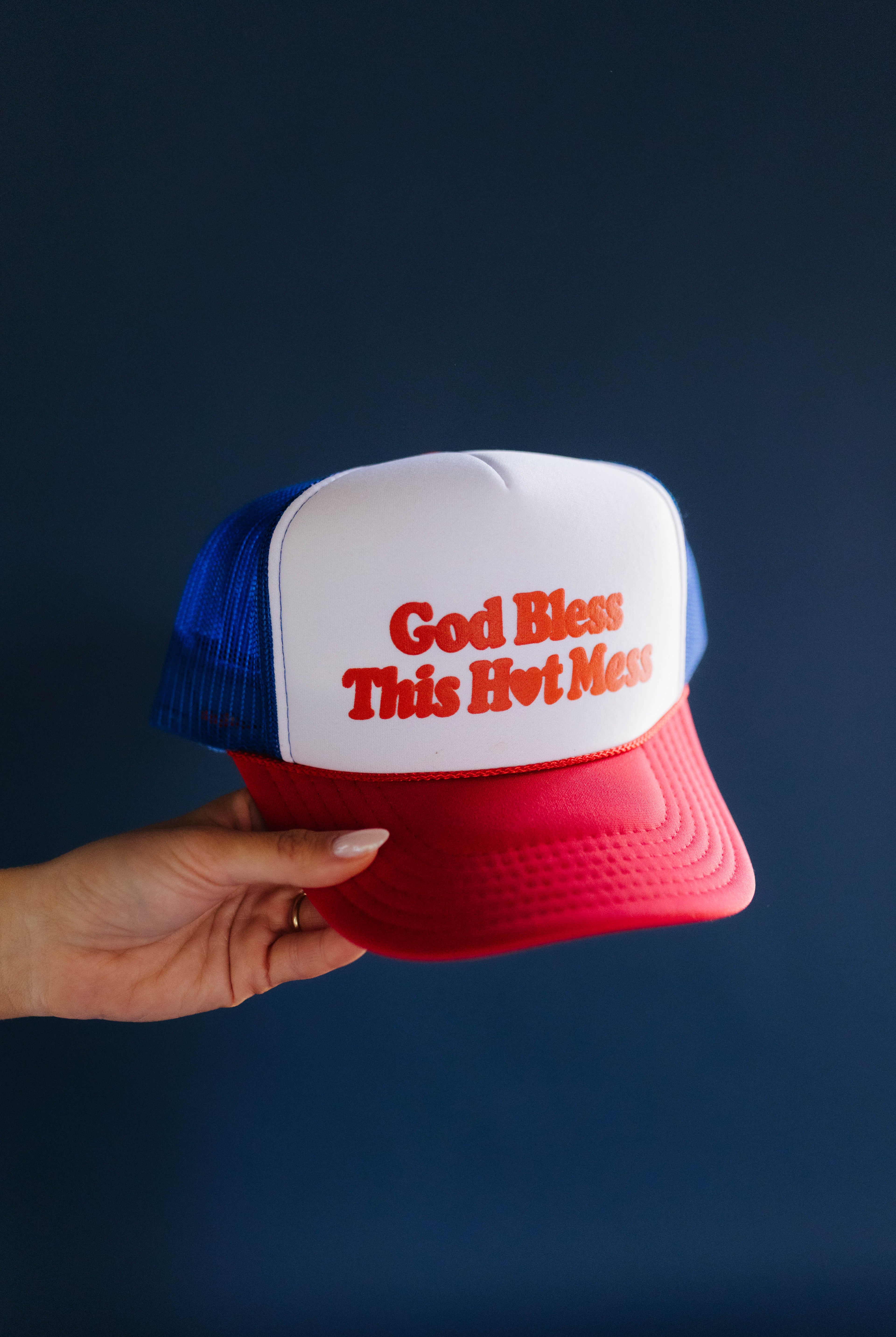 Trucker hat with 'God Bless This Hvit Moss' text held by a hand against a dark background