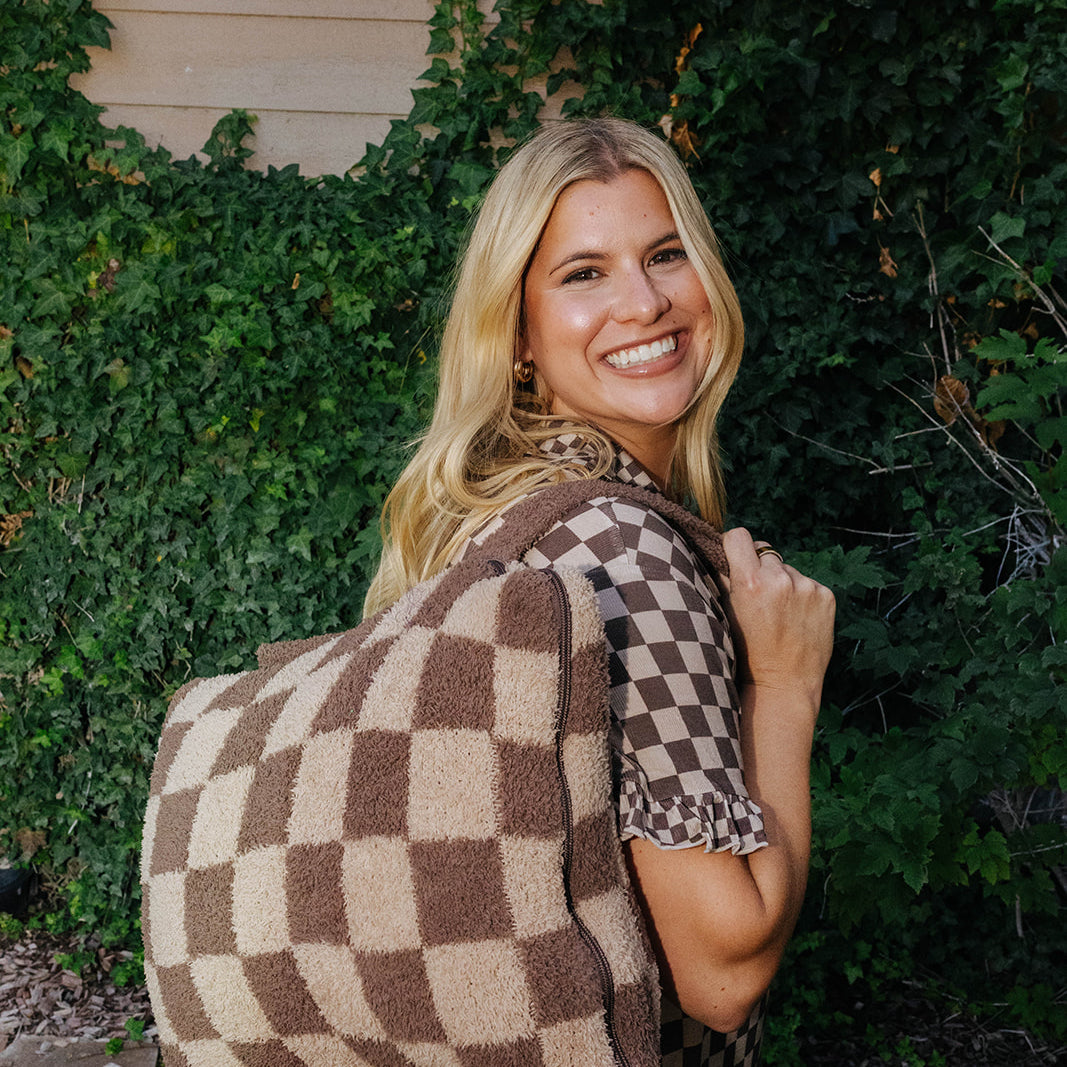 Model wearing brown check pajamas and carrying a folded brown check quillow with backpack straps.